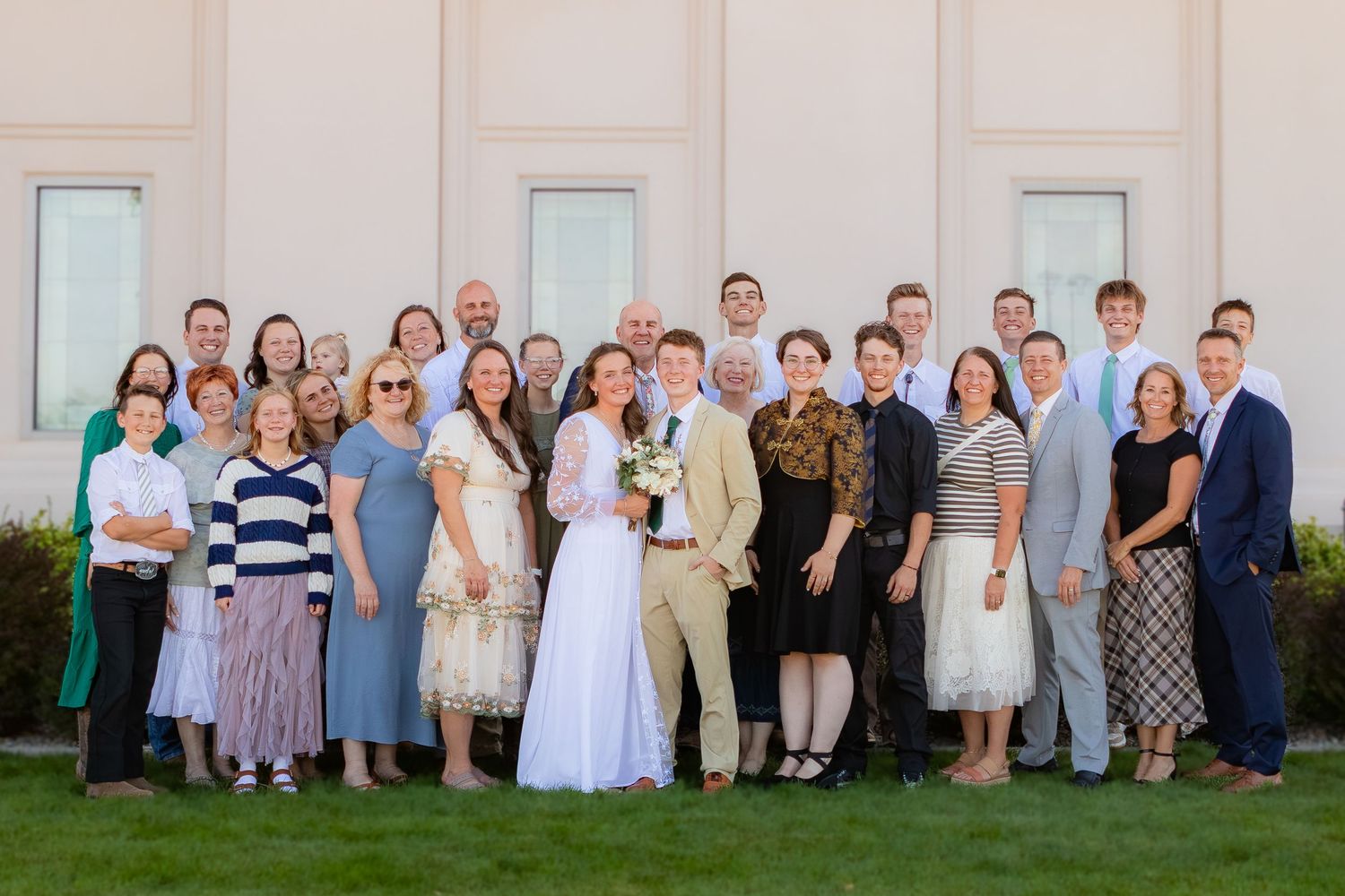 Large wedding party group photo outdoors, bride in white dress with groom and family members smiling together.