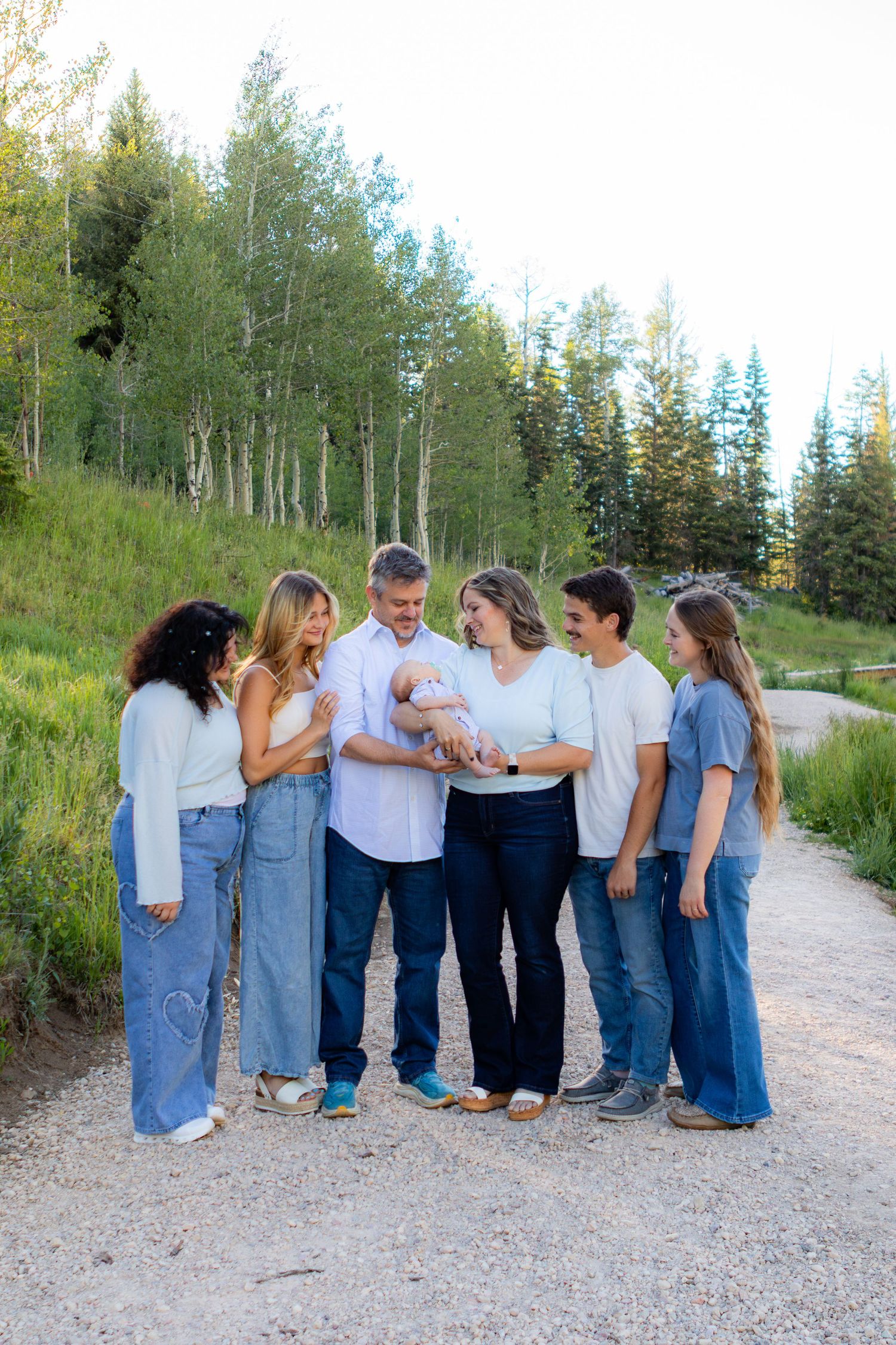 Family of six gathered on a scenic mountain path surrounded by lush green trees, admiring a newborn baby.