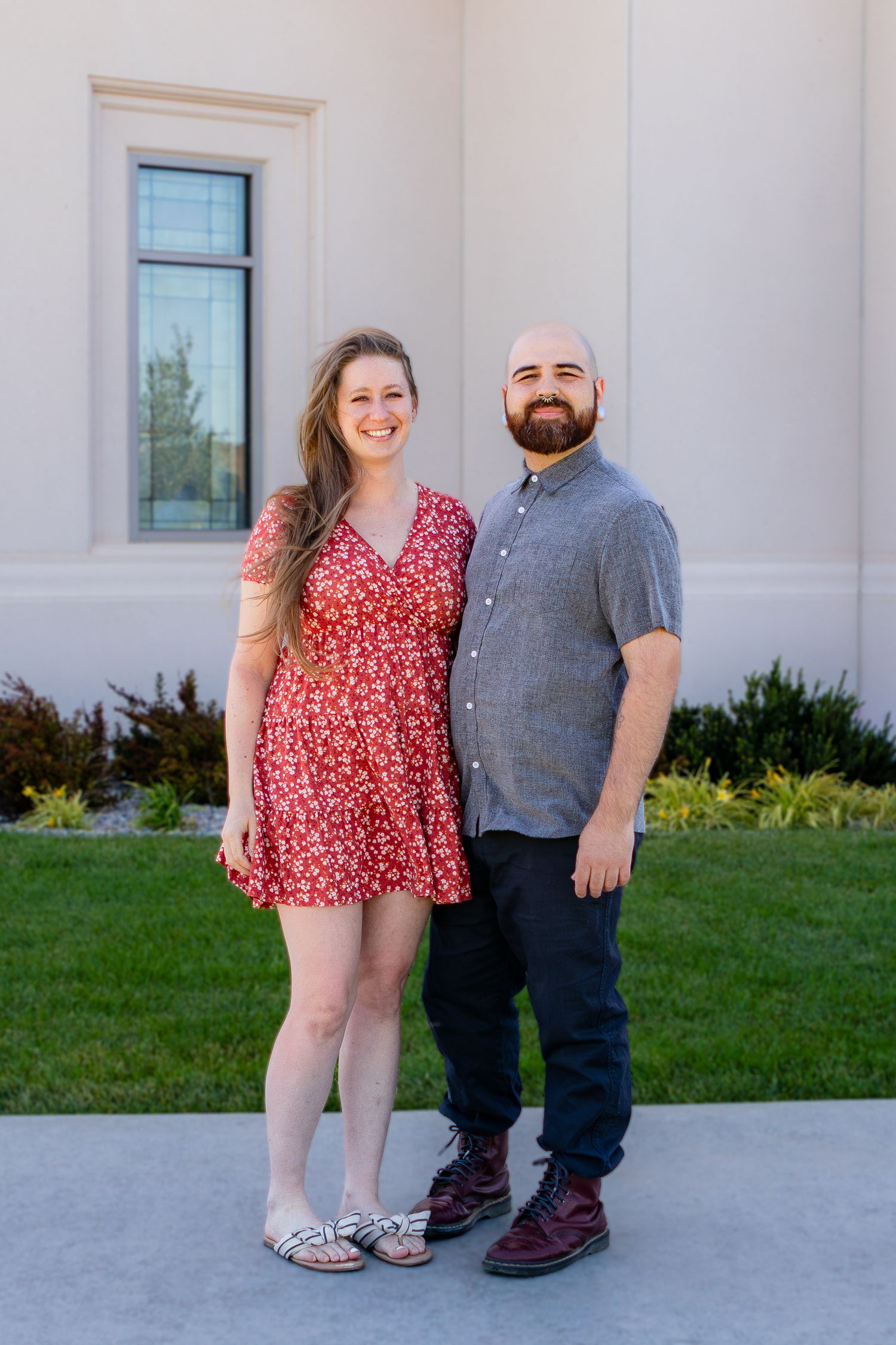 A smiling couple poses outdoors, woman in red floral dress and man in gray shirt, standing in front of a building.