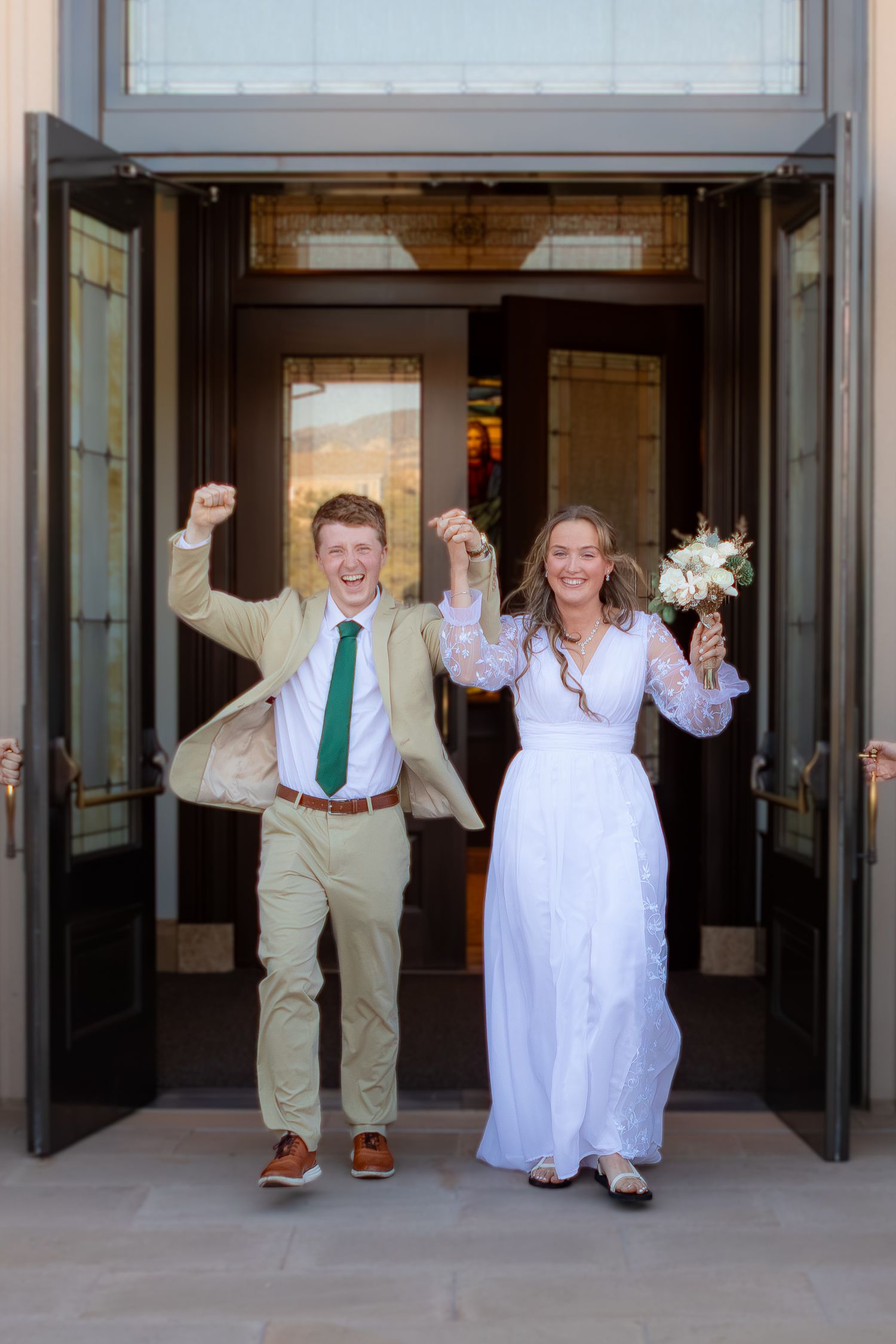 Joyful newlyweds exit church doors, bride in white lace gown holding bouquet, groom in tan suit with green tie at Cedar City UT Temple