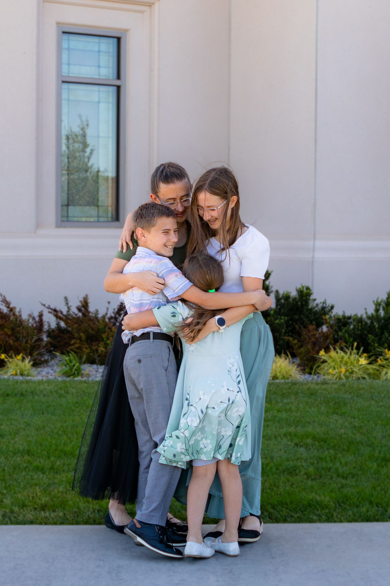 A joyful family of four shares a warm group hug outdoors near a building with green landscaping.