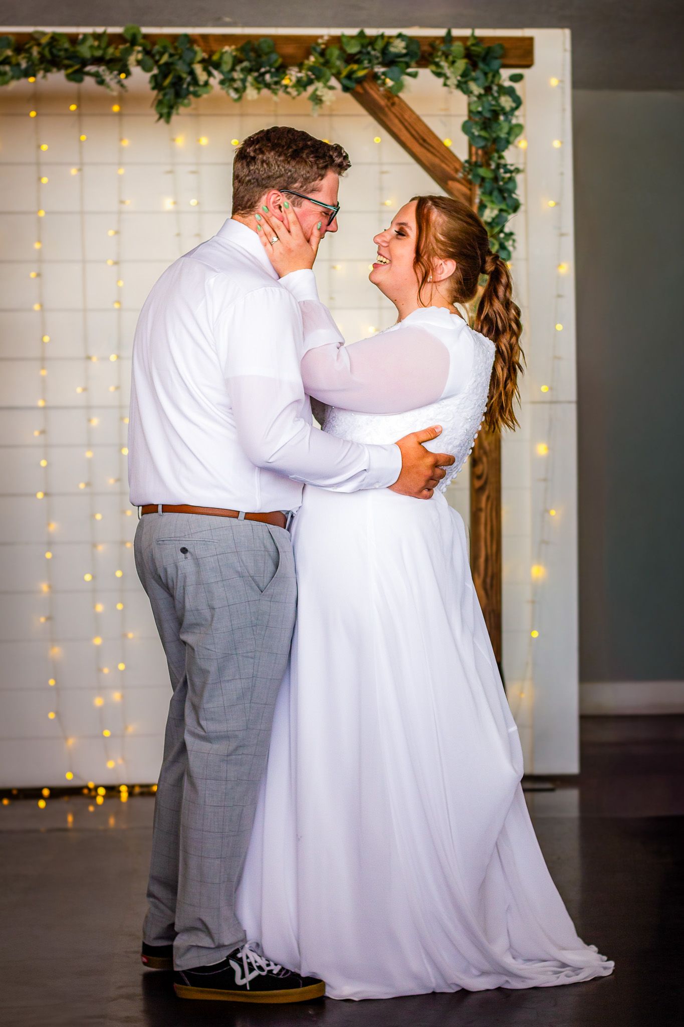 Mormon Bride and groom share a romantic embrace under a wooden arch with greenery and fairy lights at their wedding.