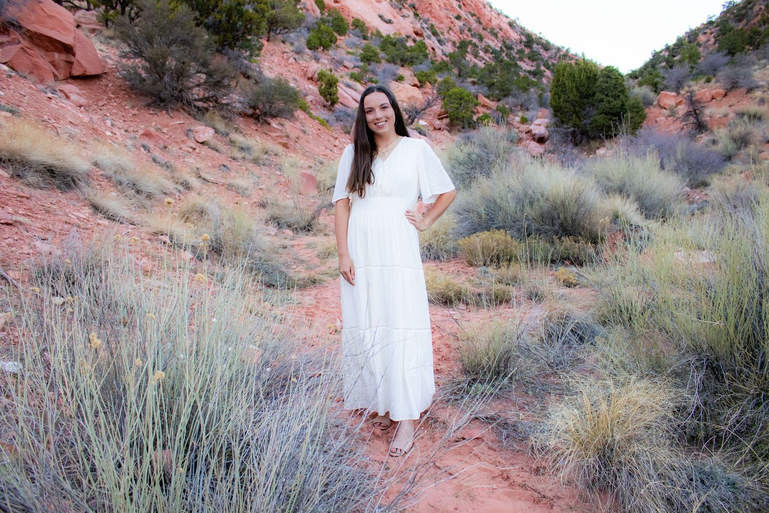 Smiling young woman in elegant white flutter-sleeve maxi dress stands on red desert trail surrounded by sage brush.