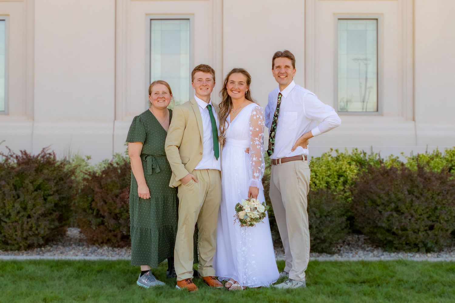 Wedding party of four smiling outdoors, bride in white gown holding bouquet, guests in formal attire near white building.