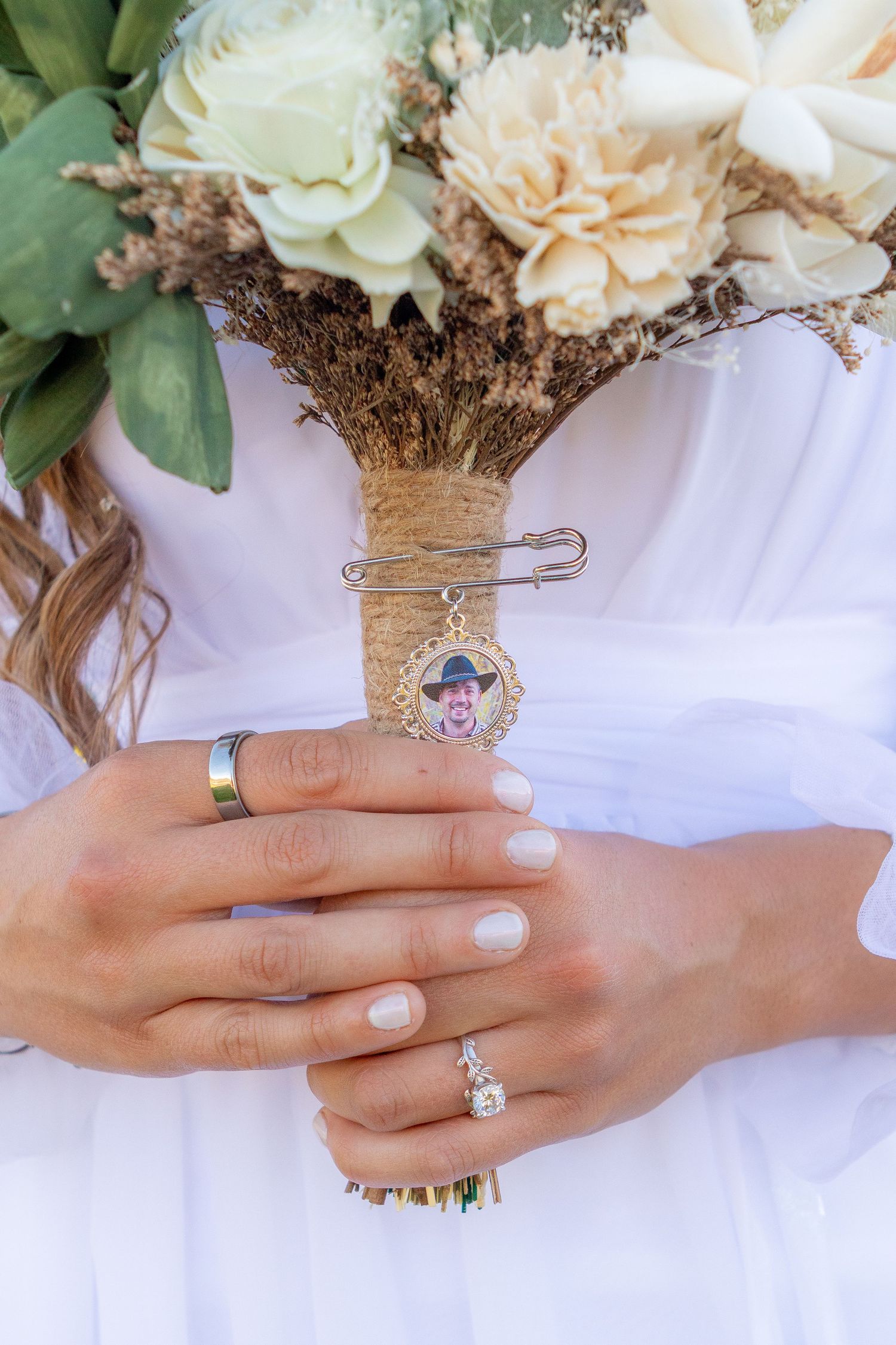 Bride holding wedding bouquet with memorial photo charm, wearing diamond engagement ring and wedding band in white dress.