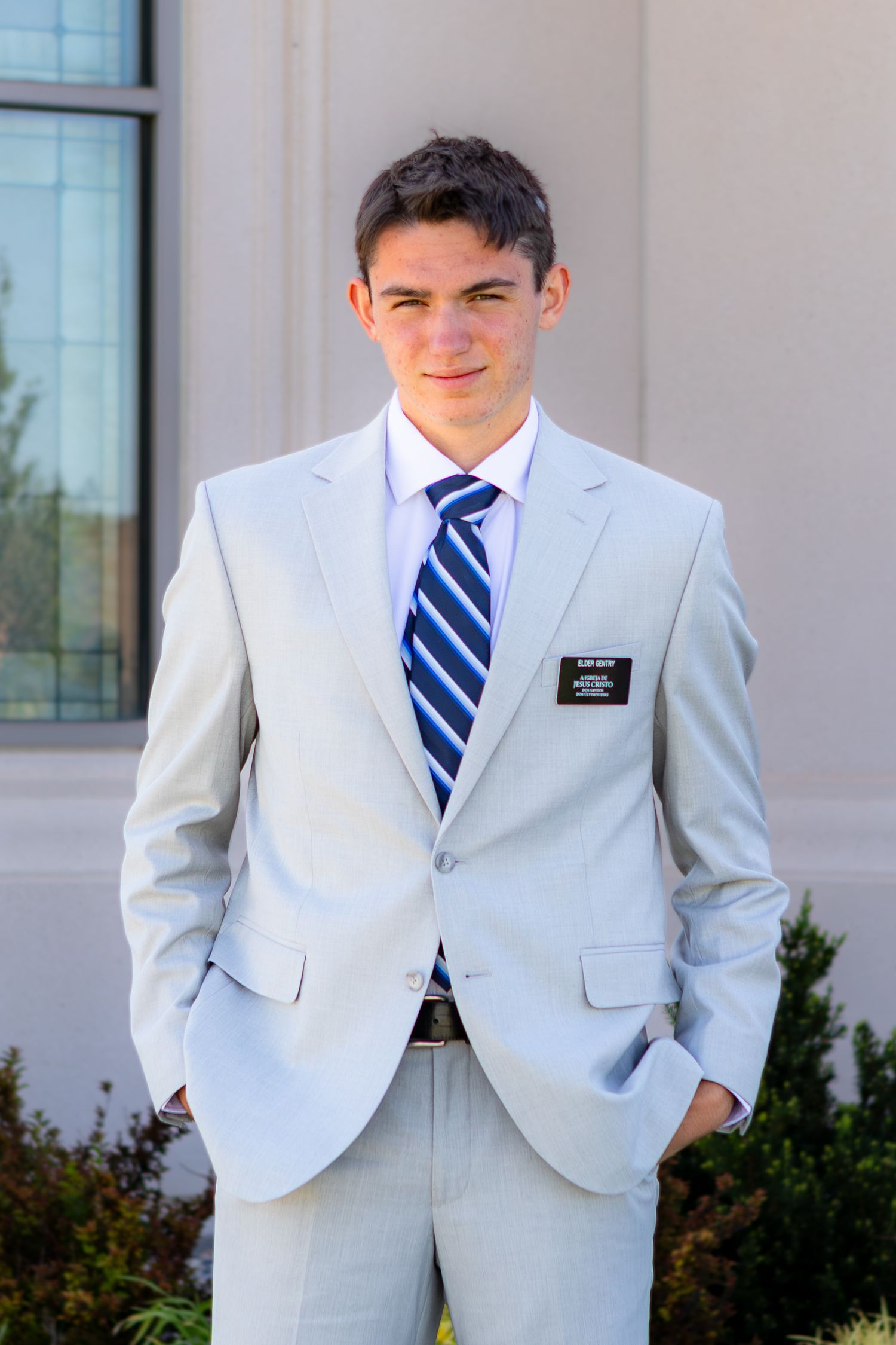 Young man in light gray suit with blue striped tie wearing a name badge, posing outdoors near a building.