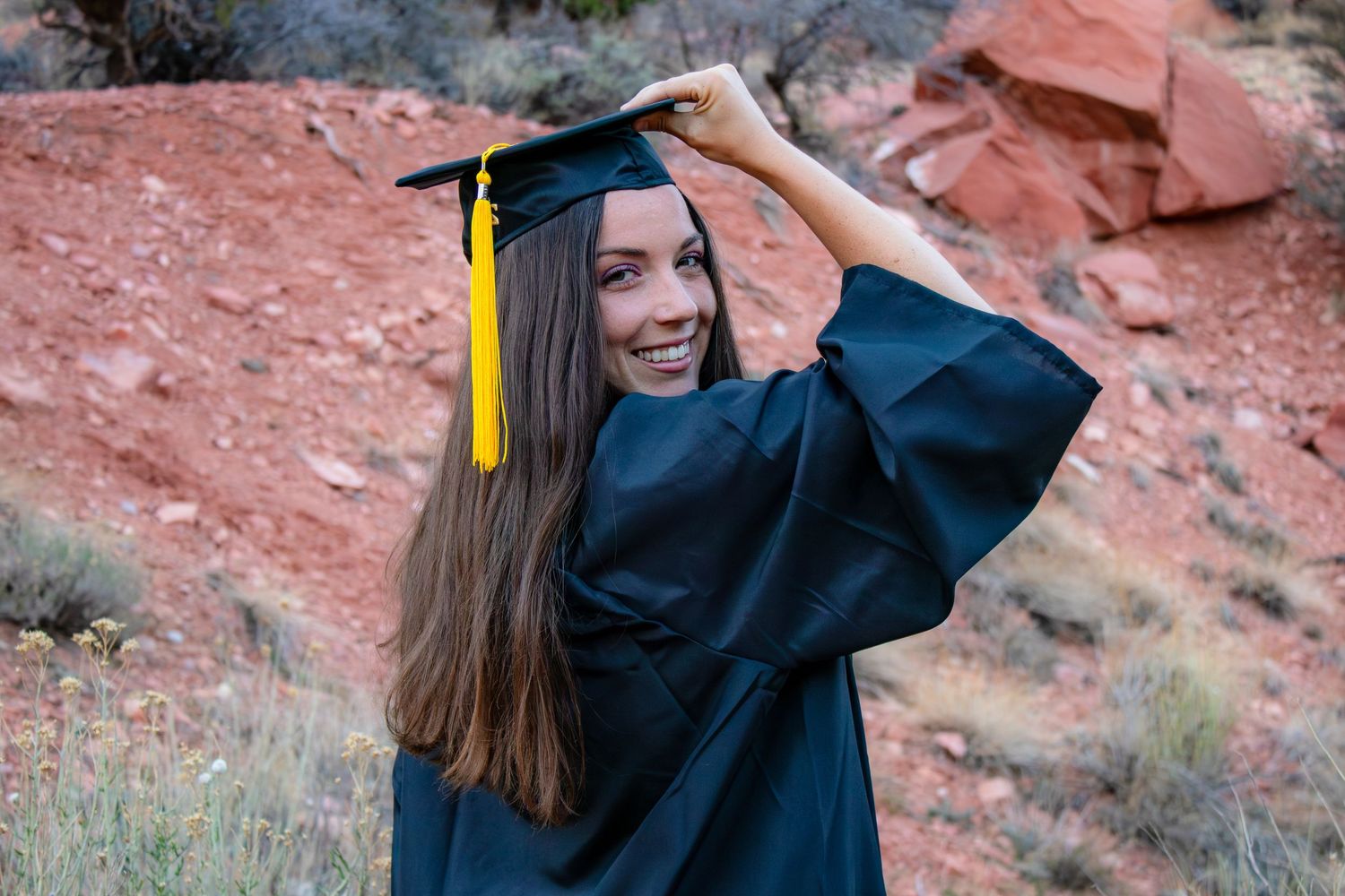 Smiling graduate in black cap and gown posing among red rock desert landscape.