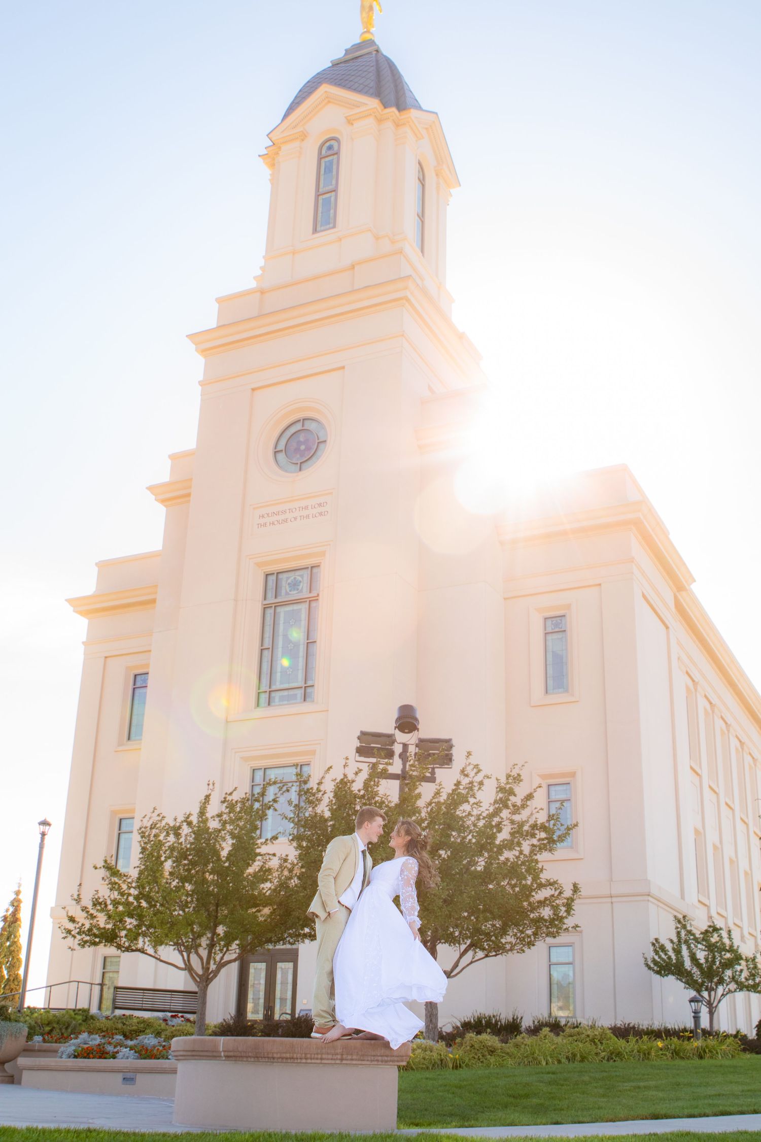 Couple embracing in white wedding attire outside a grand LDS temple with golden spire and sunburst.