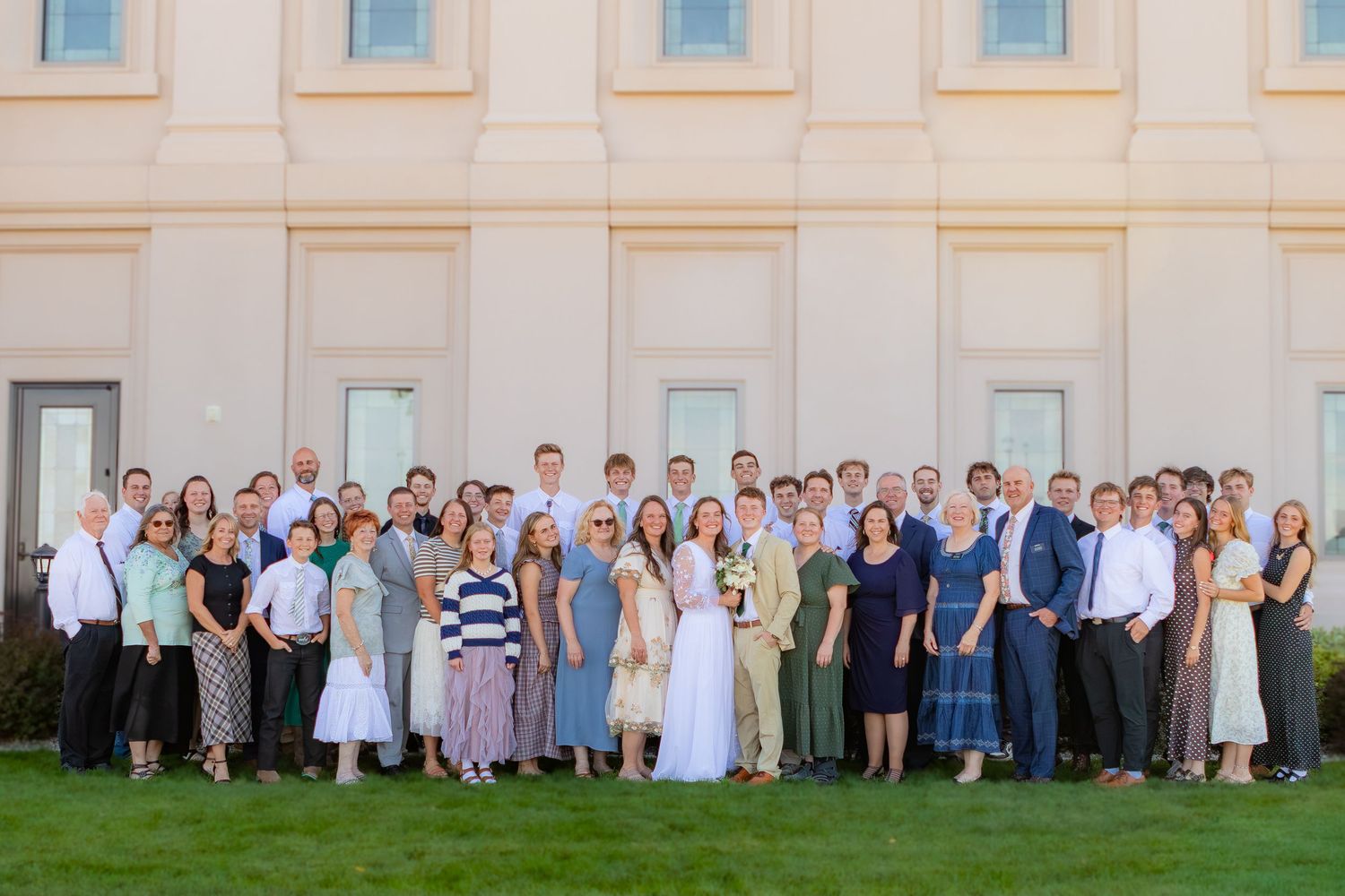 Large family group photo outside a elegant building on a sunny day, dressed in formal wedding attire on green lawn.
