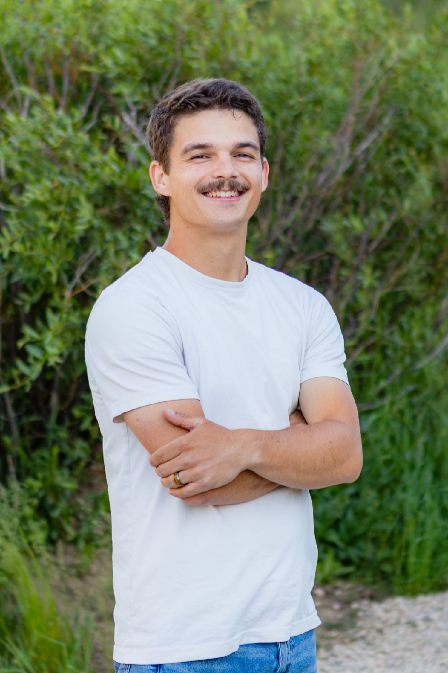 Young man with mustache smiling outdoors, wearing white t-shirt and jeans, arms crossed, with green shrubs background.