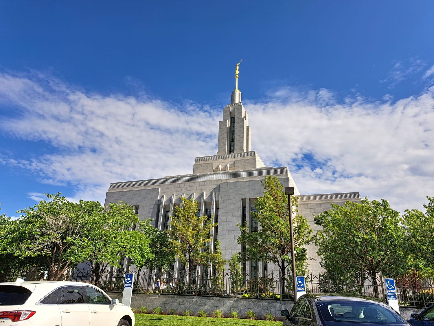 A white LDS Mormon temple with a tall spire rises against a blue sky, surrounded by green trees and a parking lot.