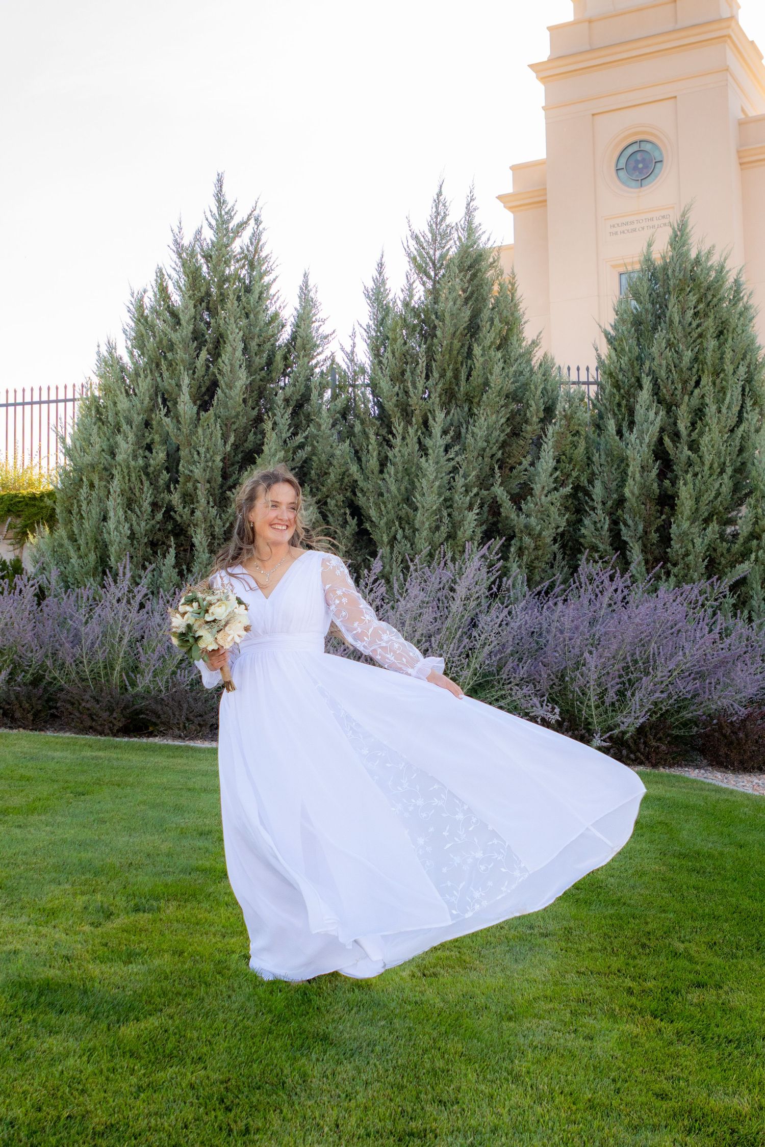 Bride in white long-sleeve gown twirling with bouquet on green lawn beside tall cypress trees and lavender bushes.