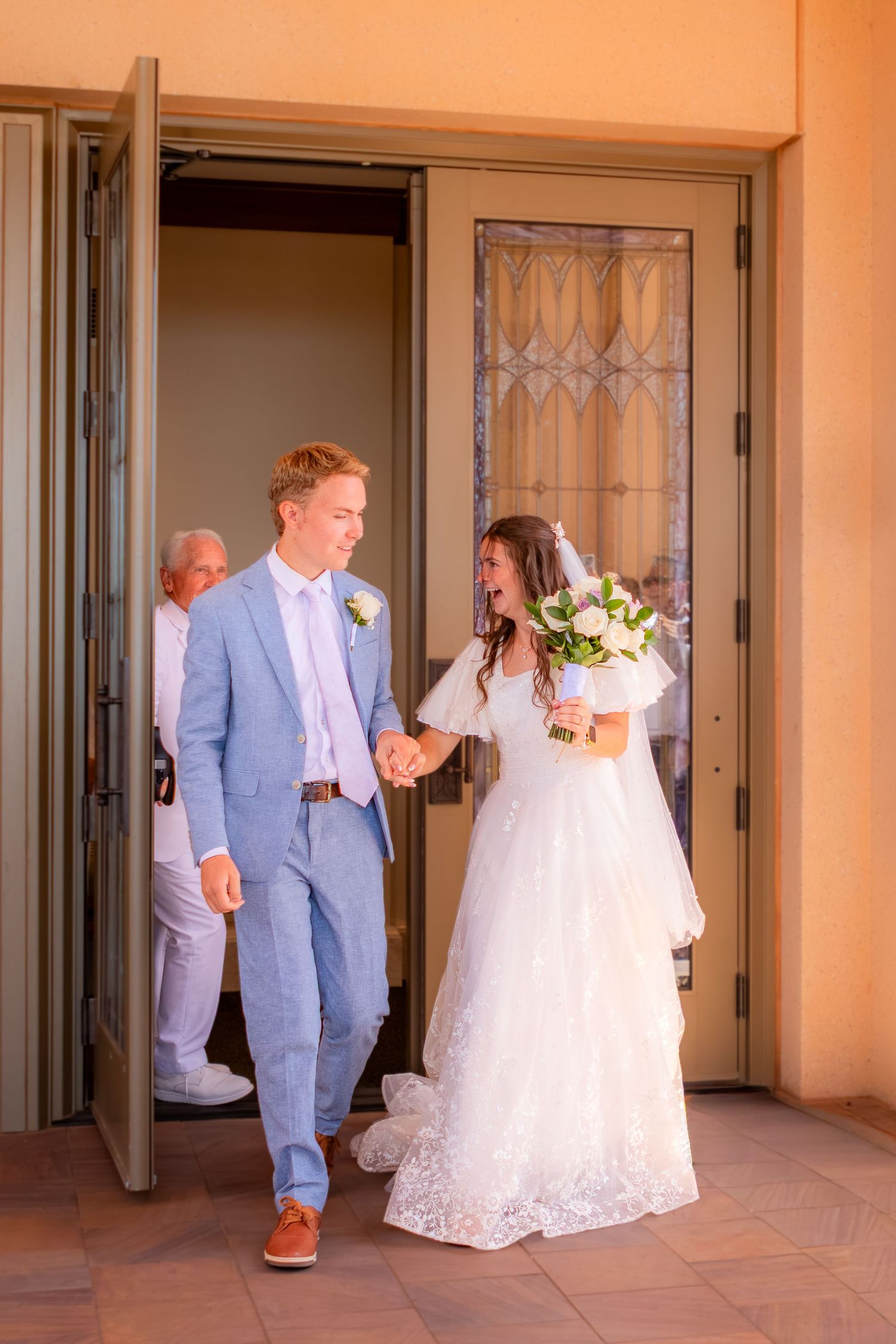 Newlywed couple exits church doors, groom in light blue suit holding bride's hand as she carries white bouquet in lace wedding gown.