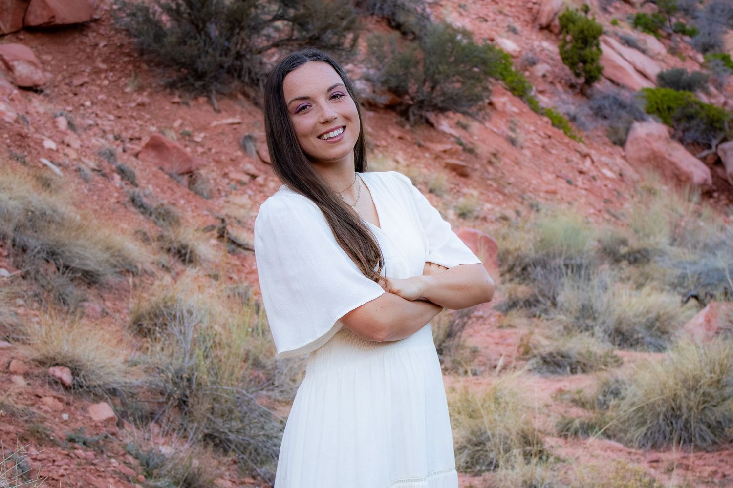 Smiling young woman in white dress with arms crossed posing before red rock desert backdrop.