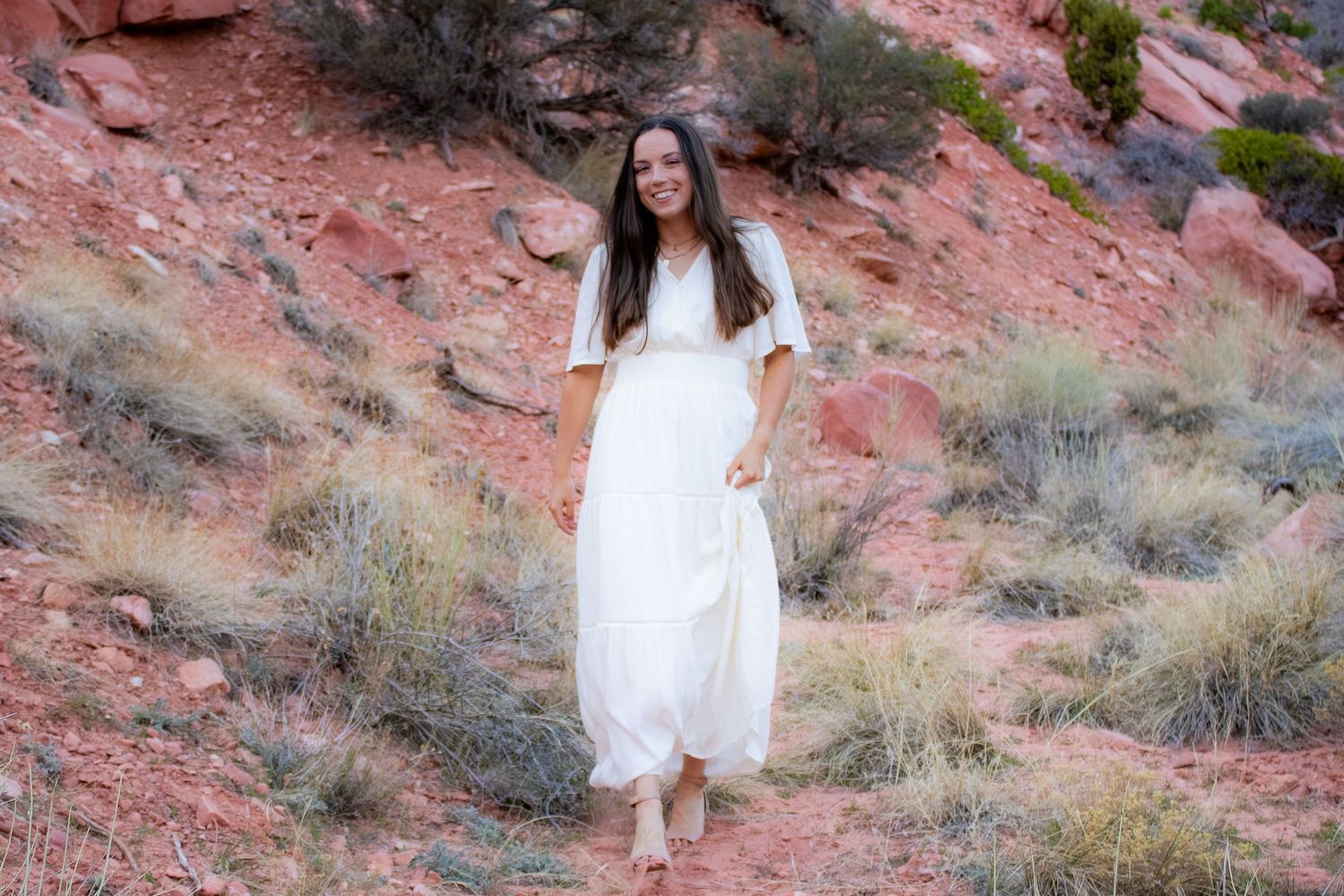 Young woman in flowing white dress walking on red rock desert trail with brush and cliffs.