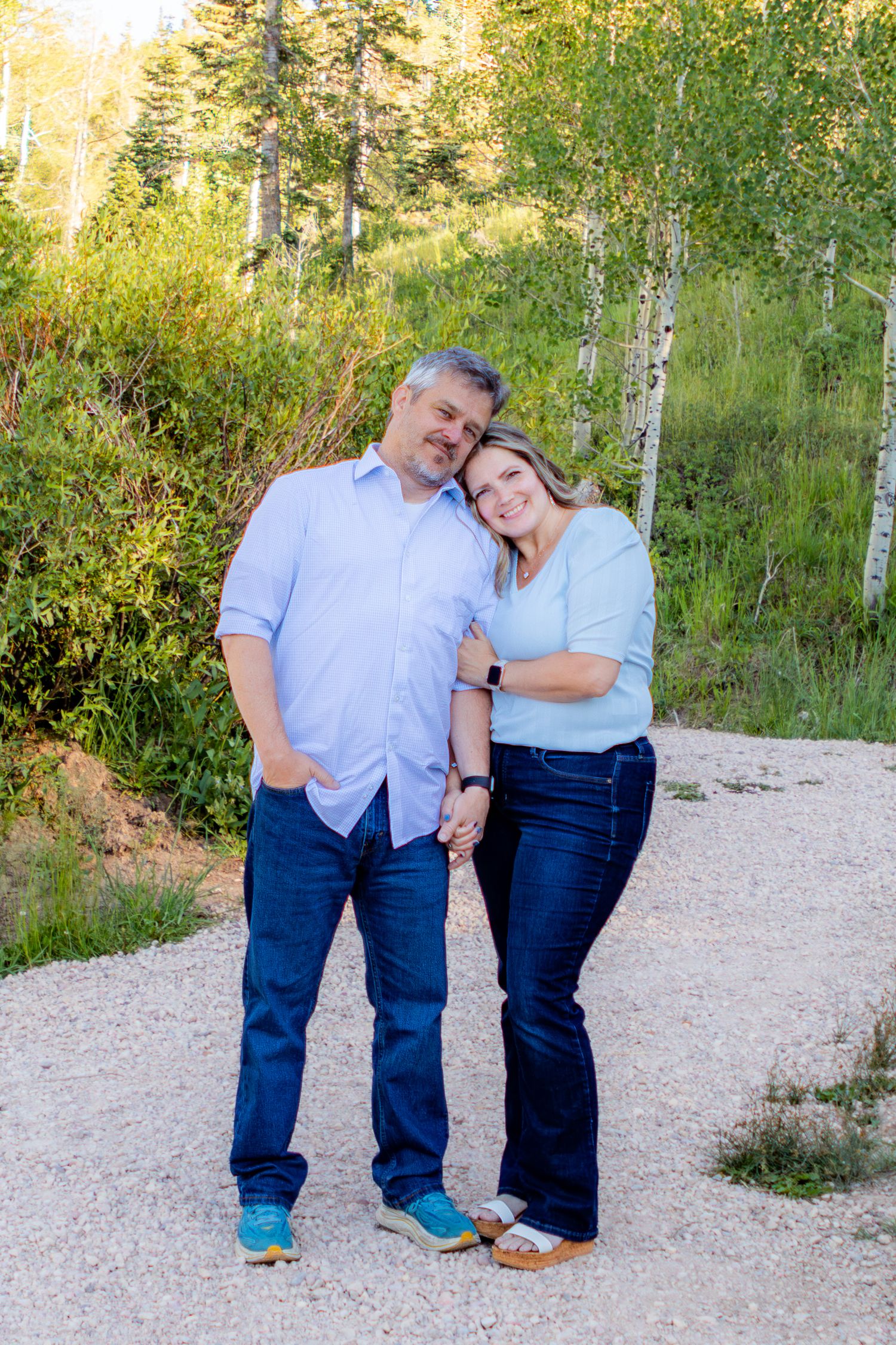A smiling couple poses hand-in-hand on a gravel path surrounded by lush green trees and aspen birches.