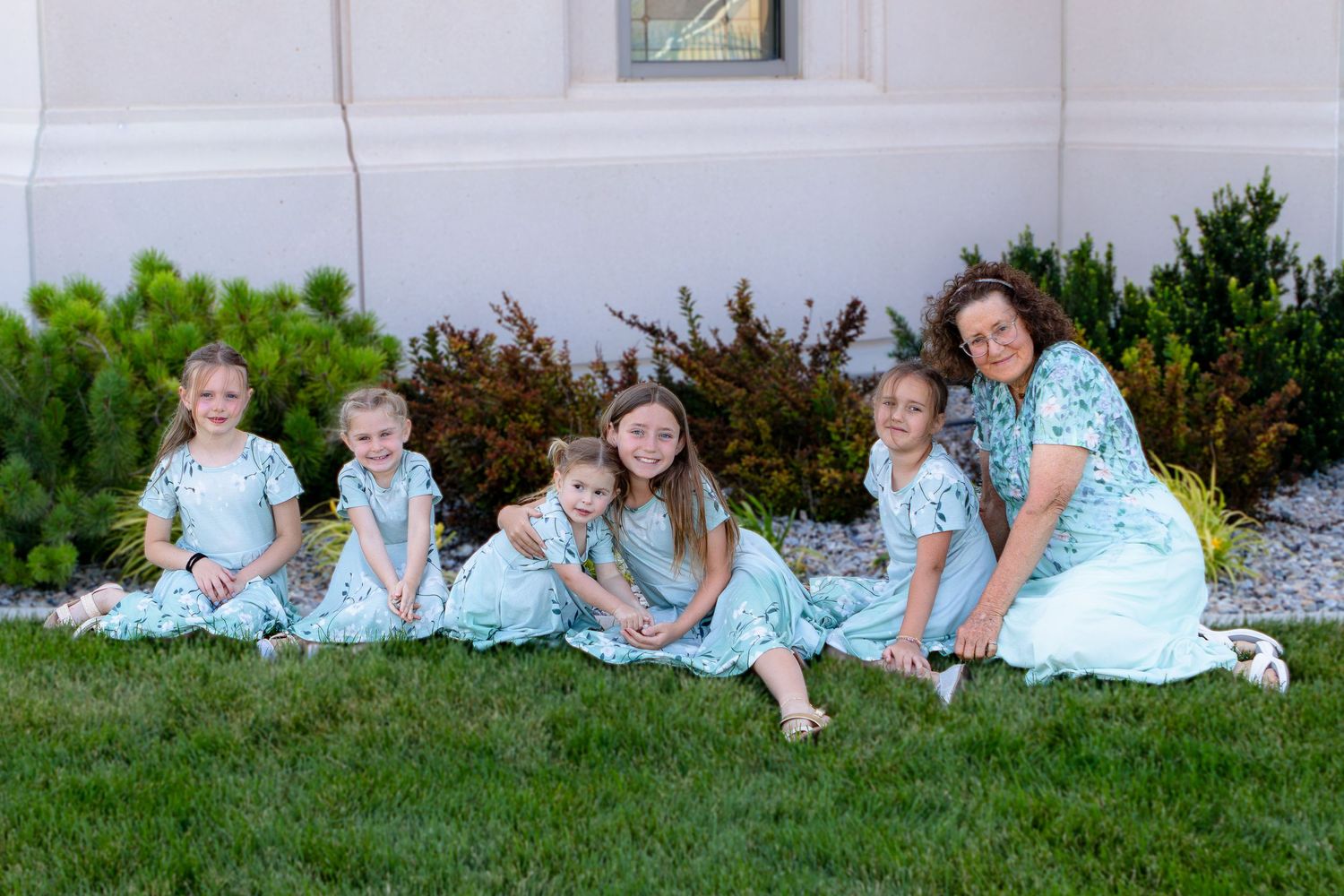 A woman and four young girls in matching teal floral dresses sitting on green grass outdoors for a family photo outside Cedar City Temple