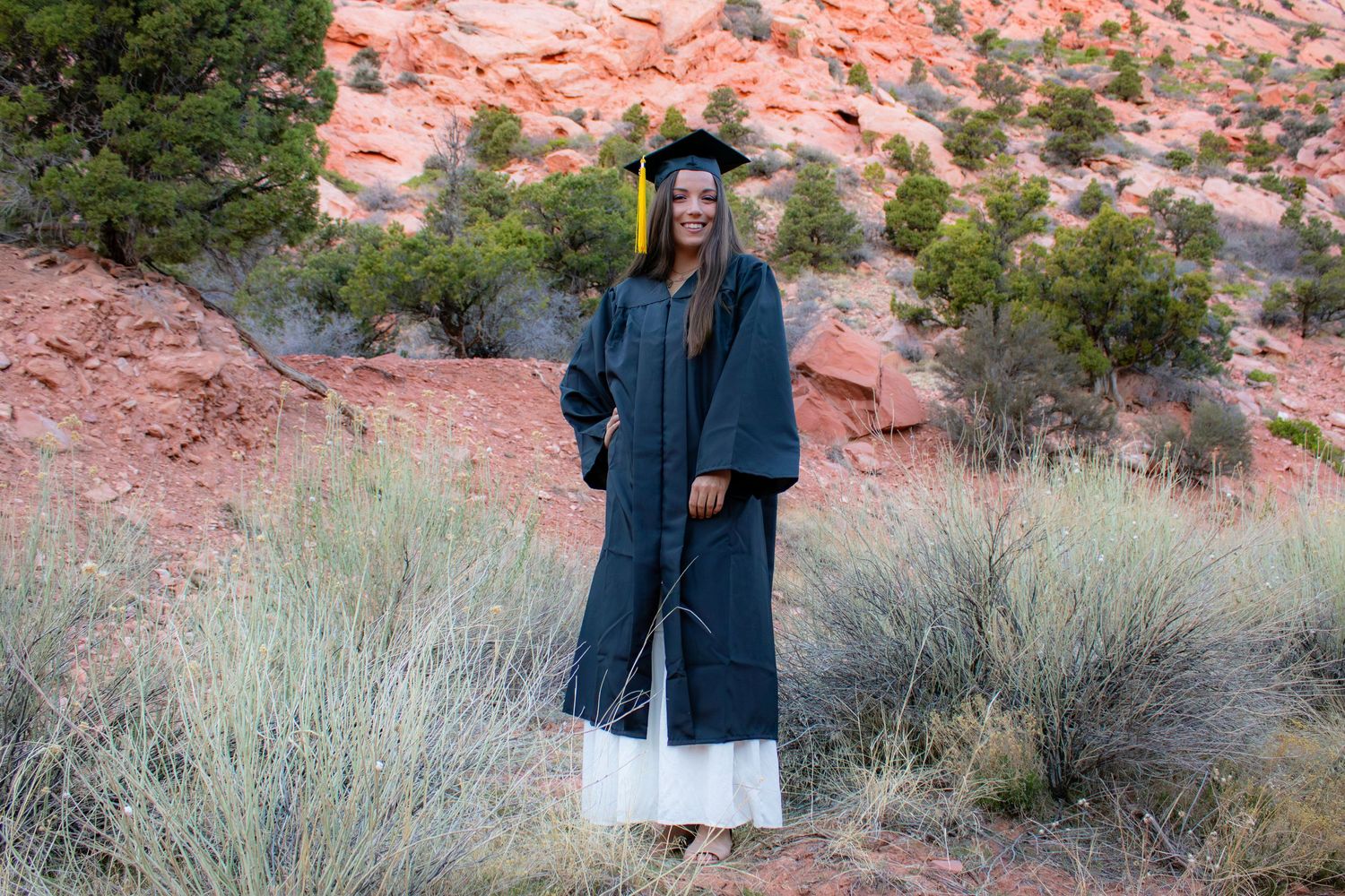 Young woman in navy graduation cap and gown smiling in front of red rock desert landscape.