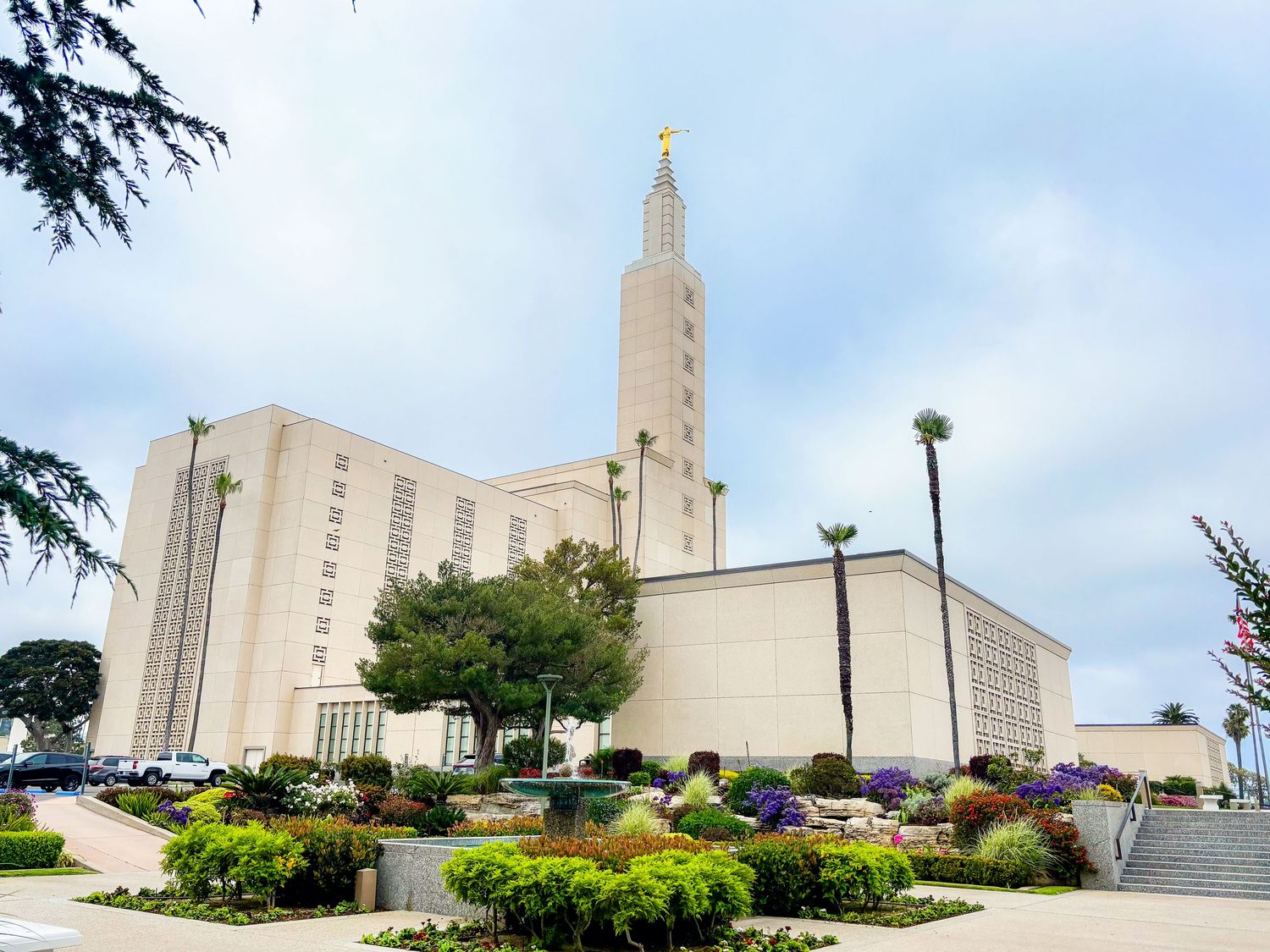 White temple with a tall central spire and gold statue, surrounded by colorful flower beds, green shrubs, and palm trees under a cloudy sky.
