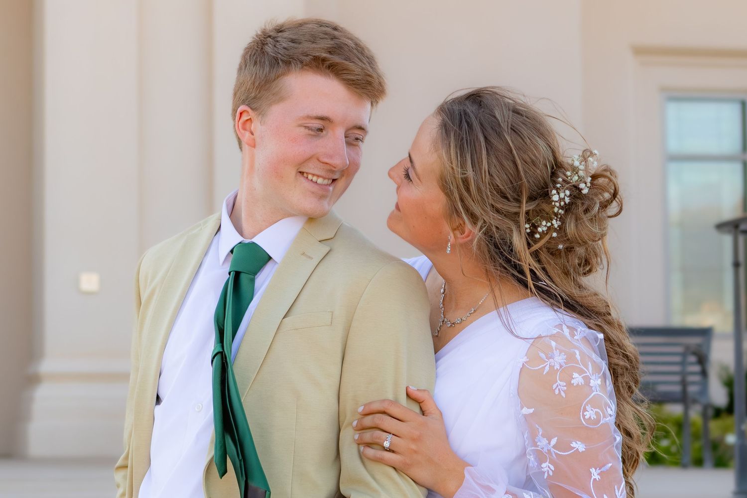 Young couple in formal attire smiling at each other, groom in tan suit with green tie, bride in white lace dress.