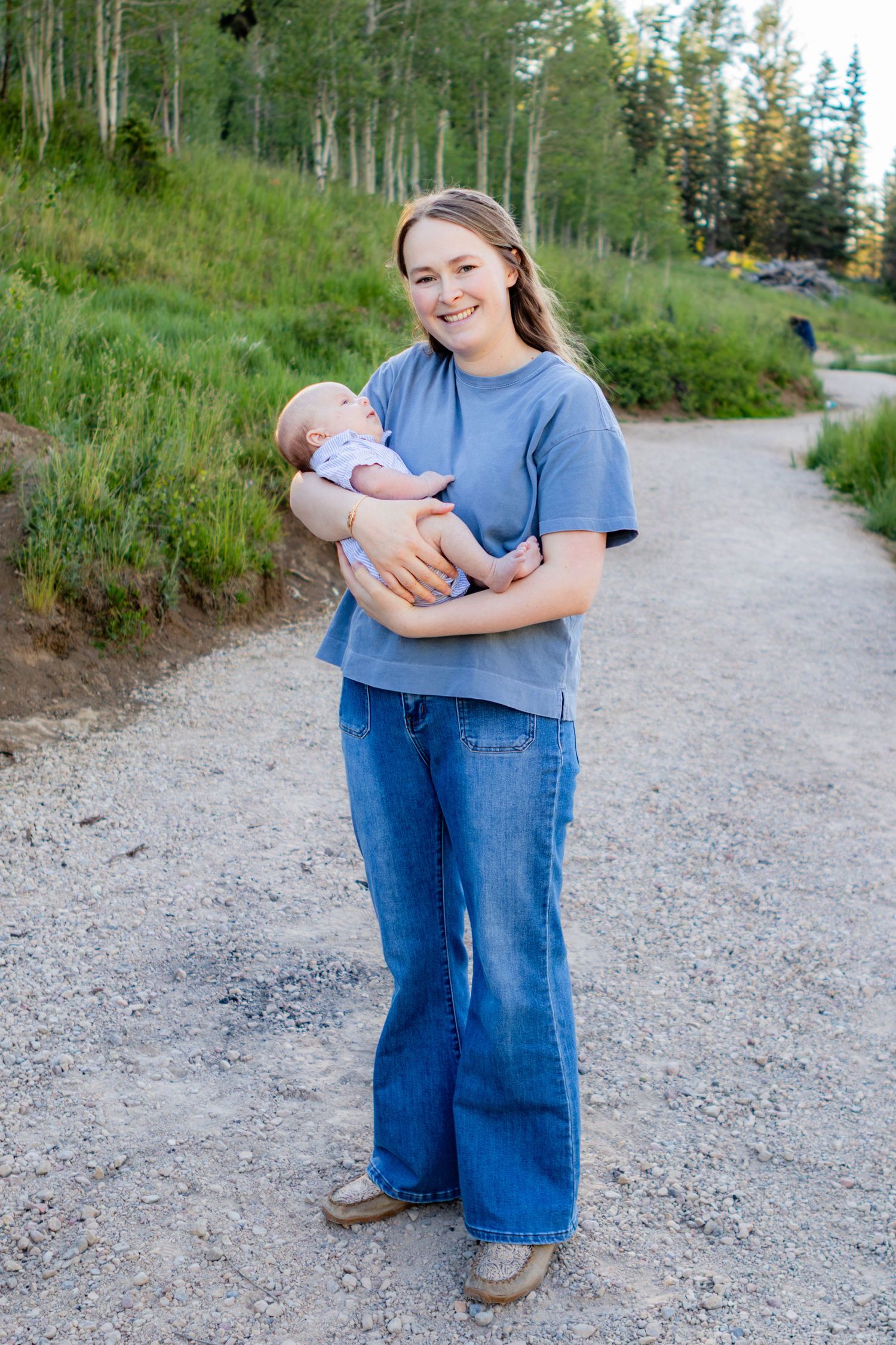 Young woman in blue t-shirt and flare jeans holds a newborn baby on a scenic gravel path surrounded by green trees.