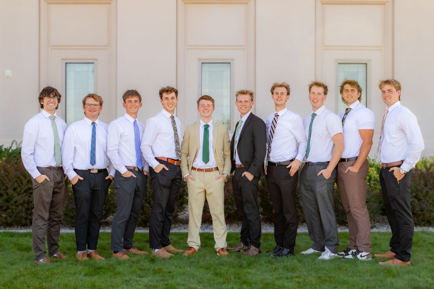 Group of ten young men in dress shirts and ties posing outdoors in front of a building on a sunny day.