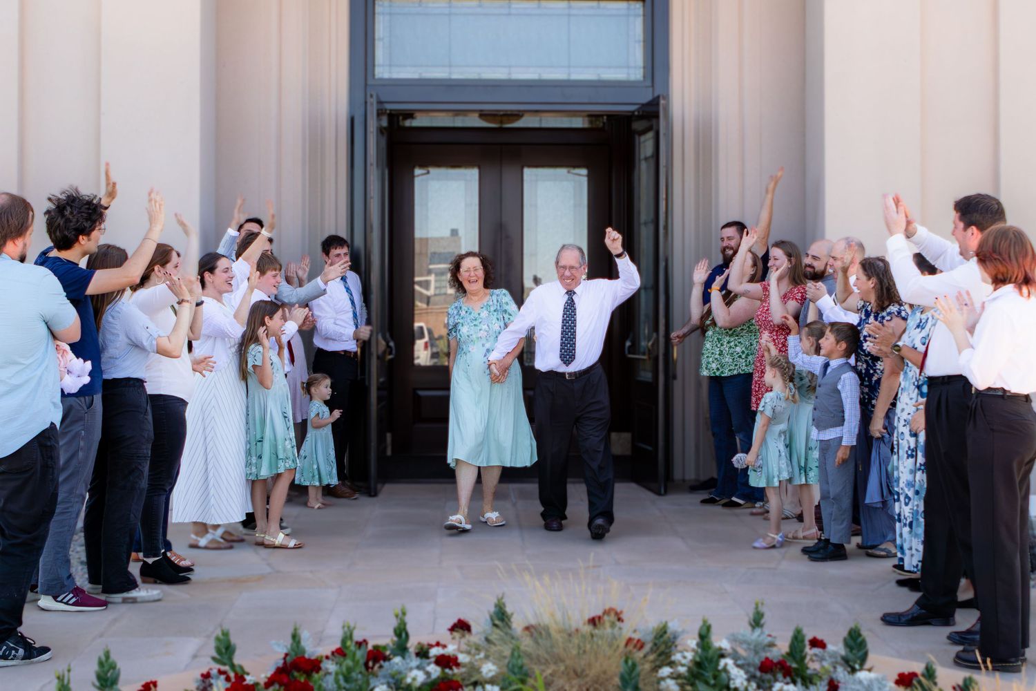 Joyful couple exits wedding venue as guests celebrate with raised hands on sunny day.
