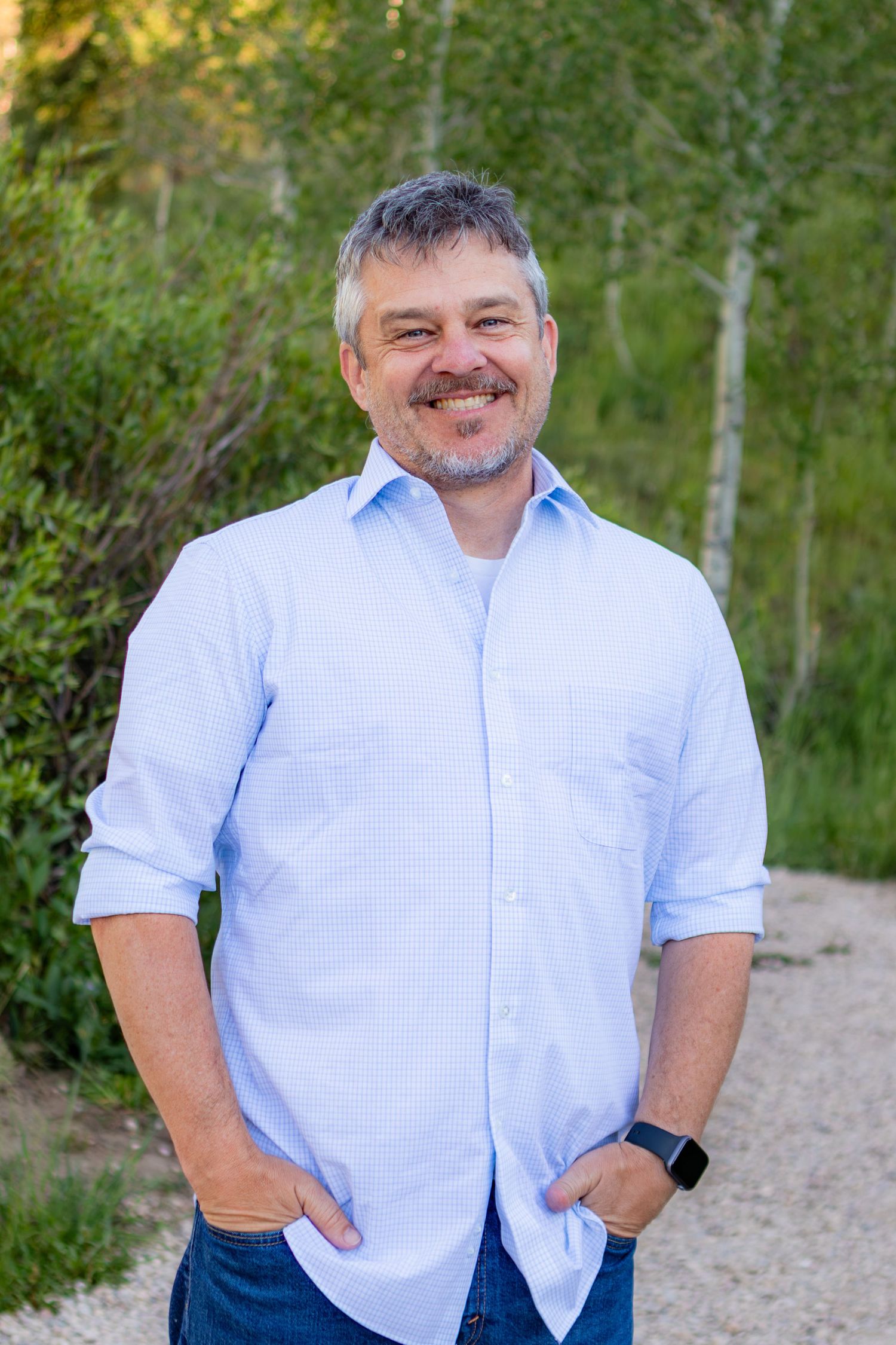 Smiling middle-aged man with gray hair and beard wearing a light blue shirt, standing on a gravel path outdoors.