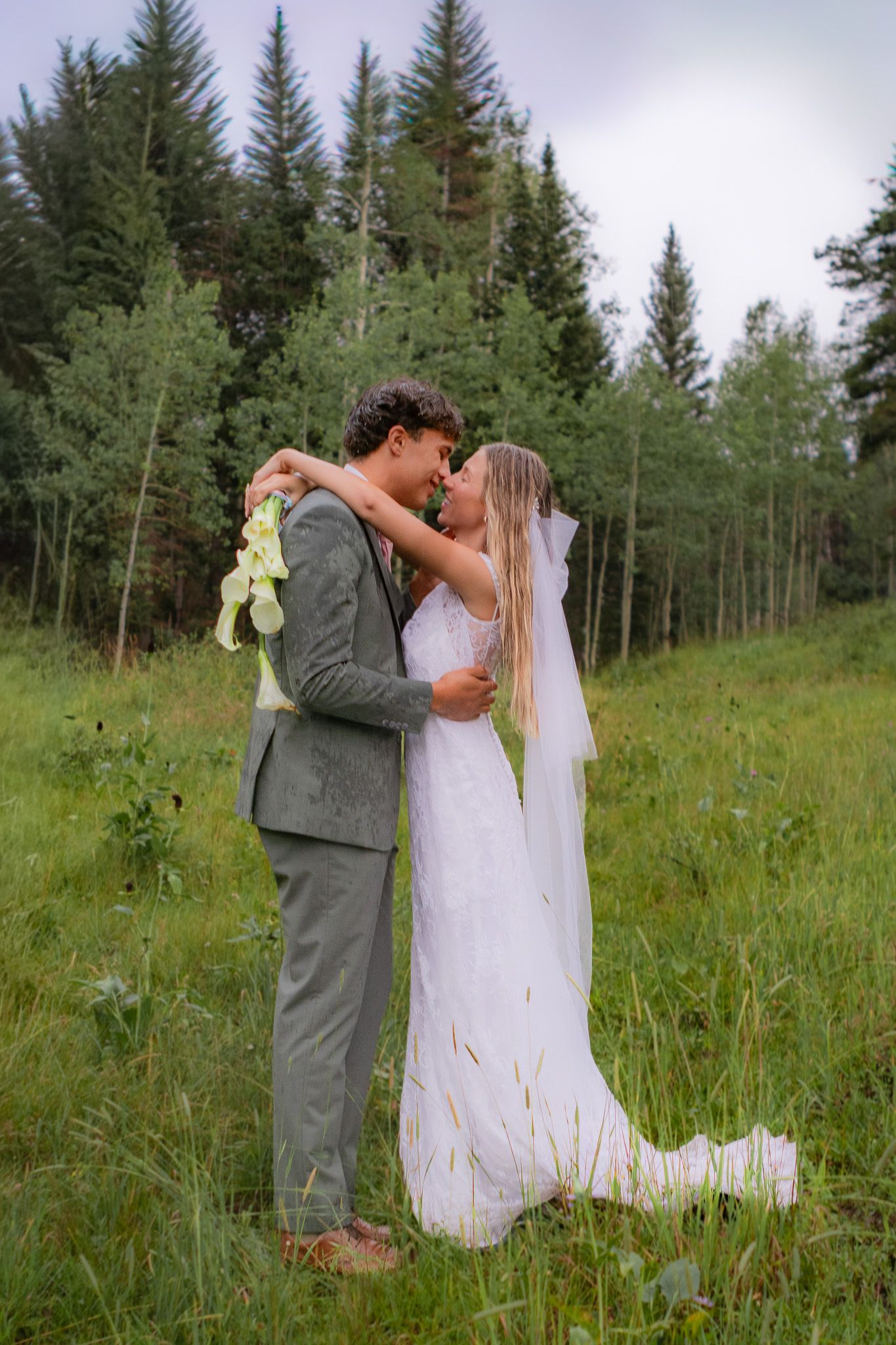 Bride and groom share a romantic moment in a lush green meadow surrounded by evergreen trees on their wedding day.