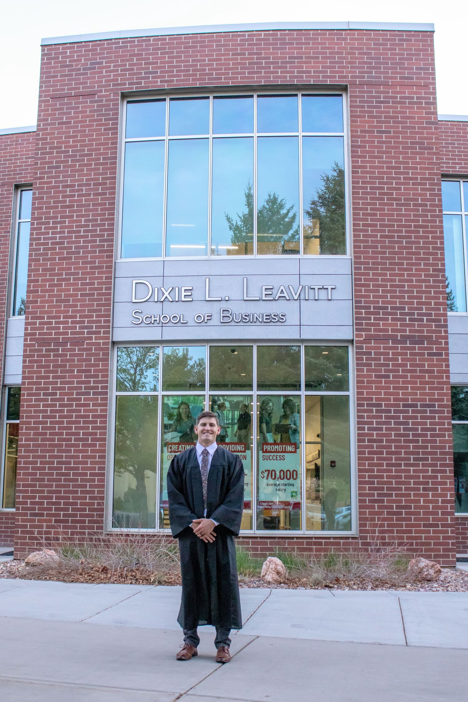 Graduate in cap and gown stands before Dixie L. Leavitt School of Business brick building at commencement.