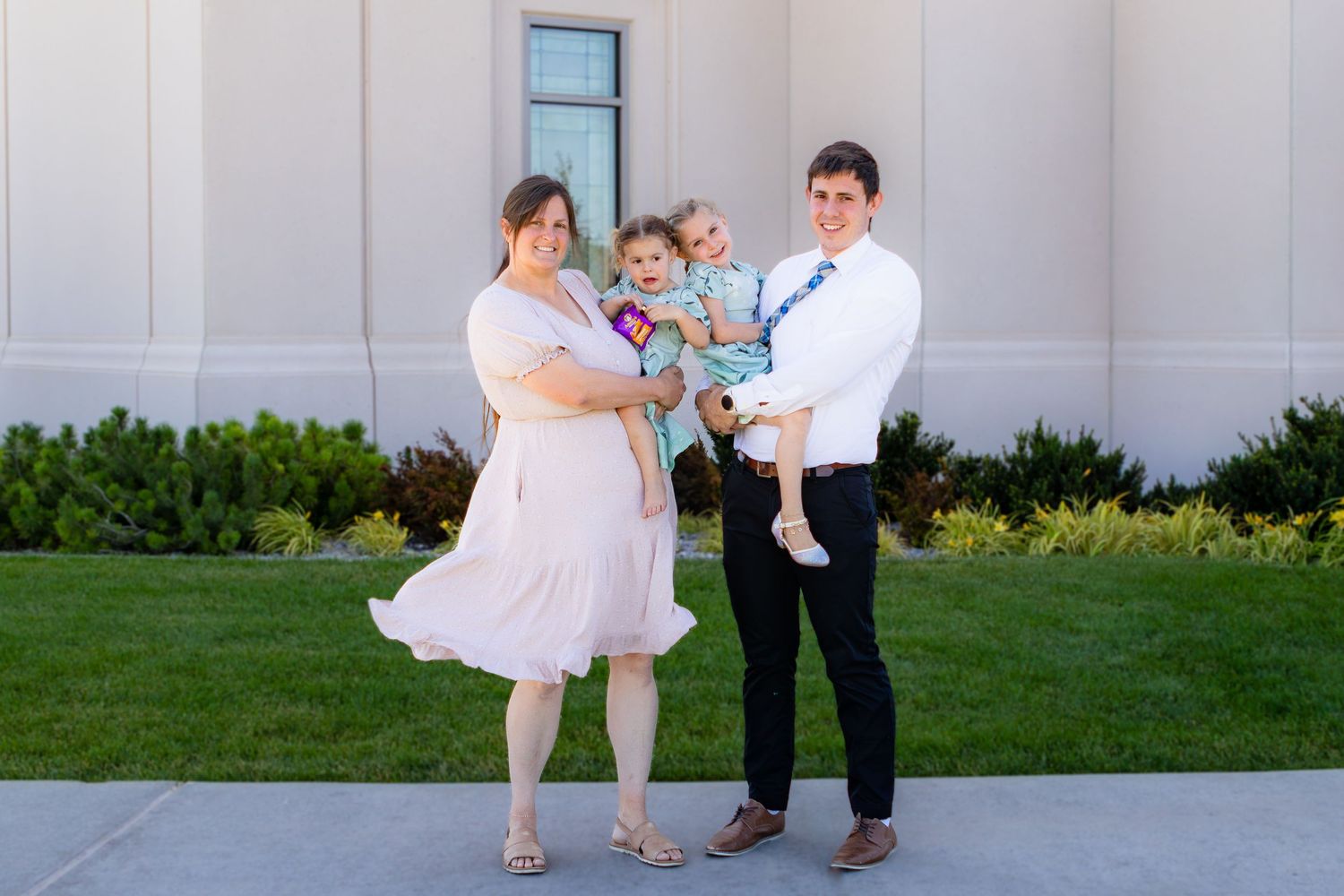 Family of four posing outdoors near the Cedar City Temple, parents holding toddlers, dressed in formal attire on green lawn.
