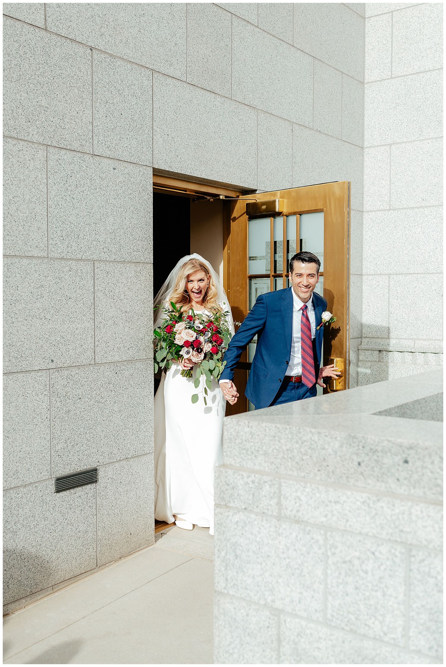 Joyful bride and groom exit church doors, bride holding red bouquet, groom in navy suit, sharing a happy moment.