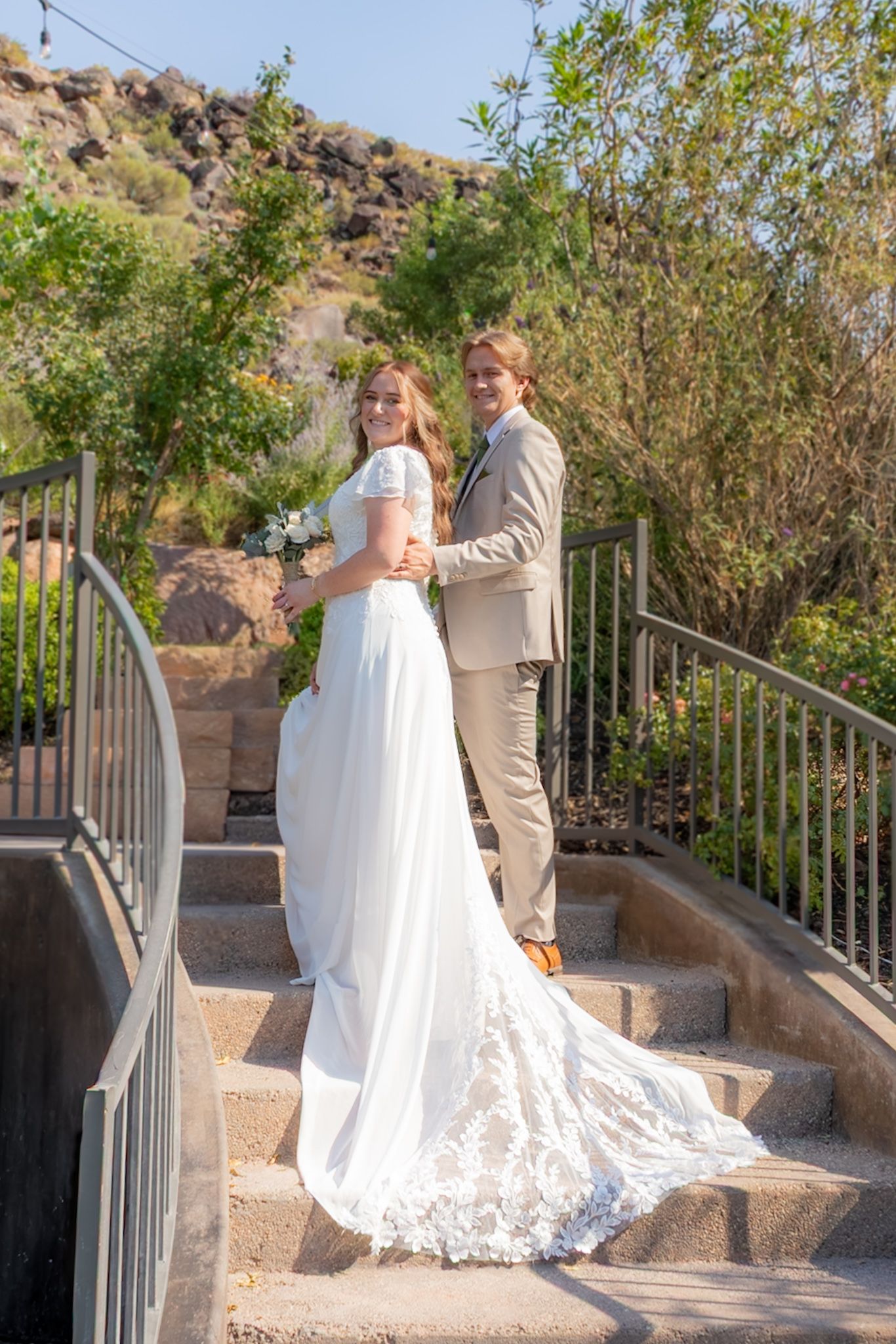 Bride and groom pose on outdoor stone steps surrounded by lush greenery and rocky hills on their wedding day.