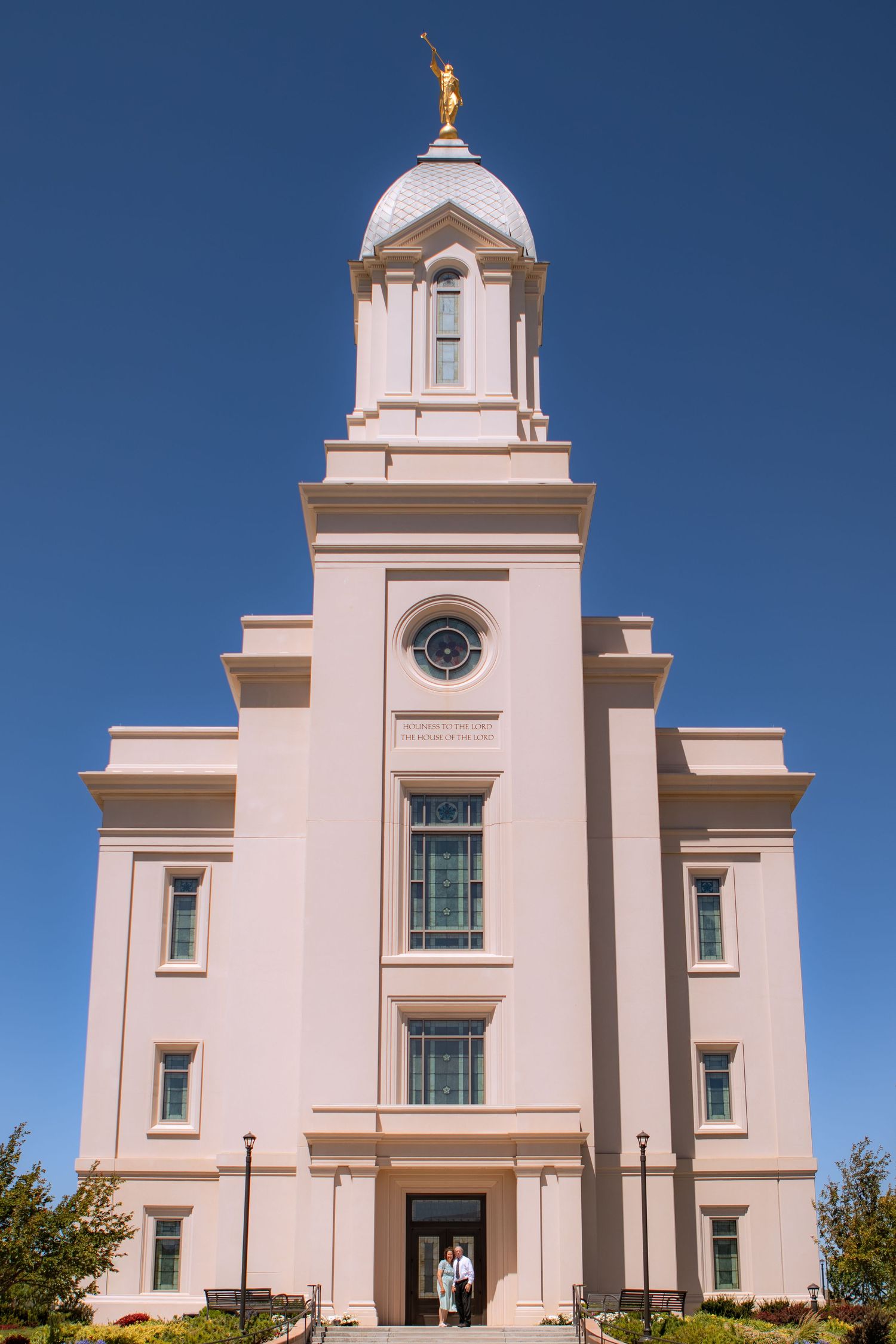 White LDS Mormon temple with gold angel Moroni statue on spire against clear blue sky, couple standing at entrance.