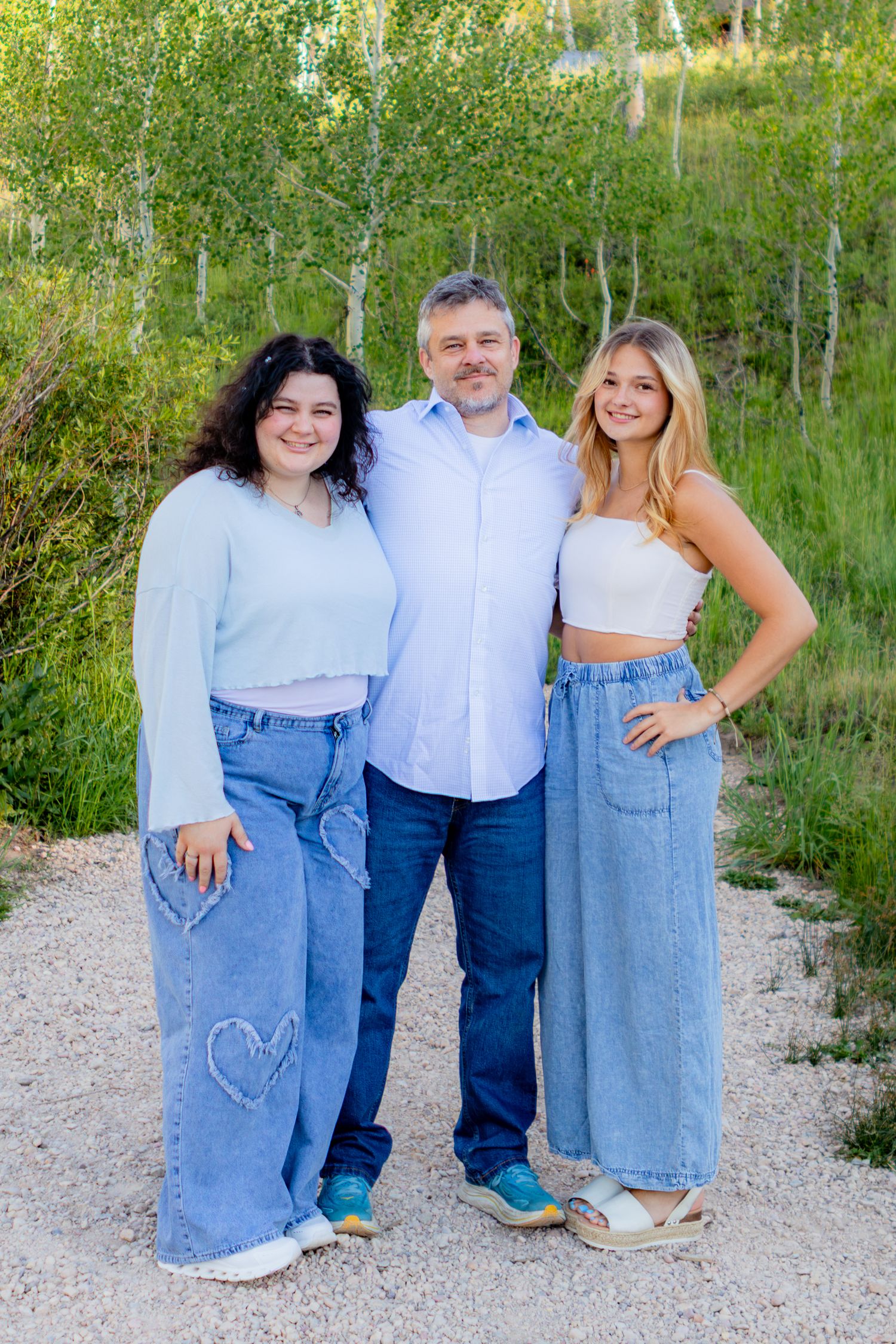 A man and two young women smile together outdoors on a gravel path surrounded by lush green trees in Cedar City UT