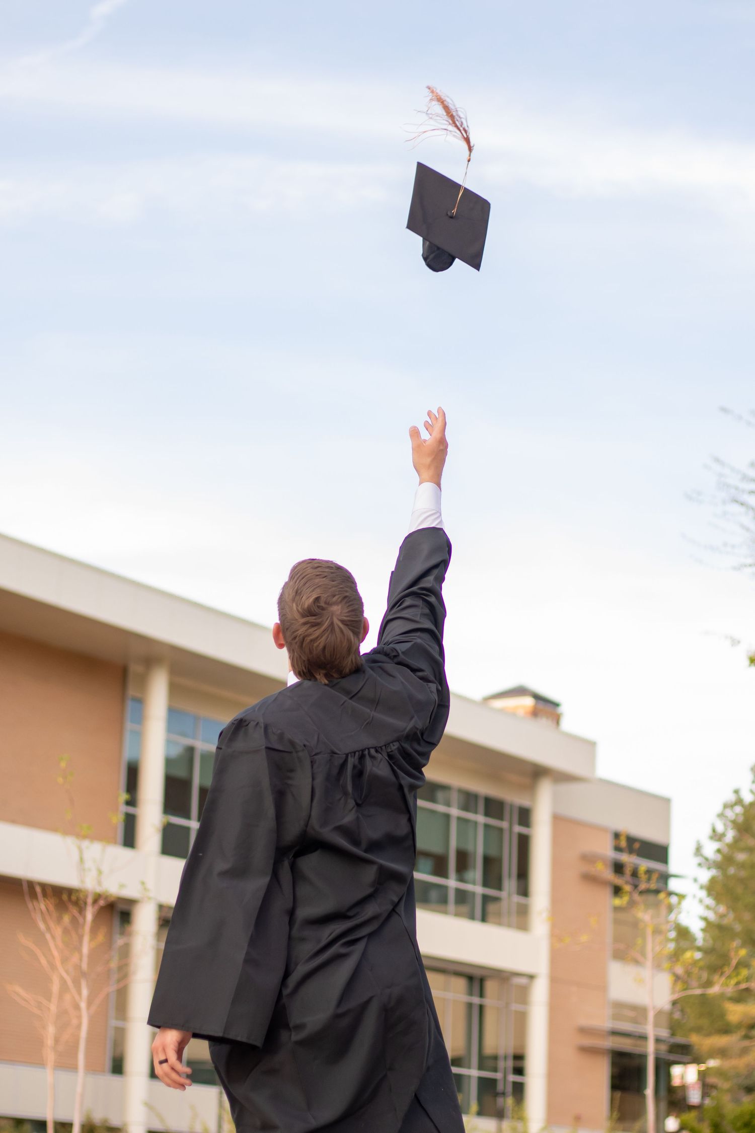 Graduate in black gown tossing mortarboard cap into sky outdoors near campus building on graduation day.
