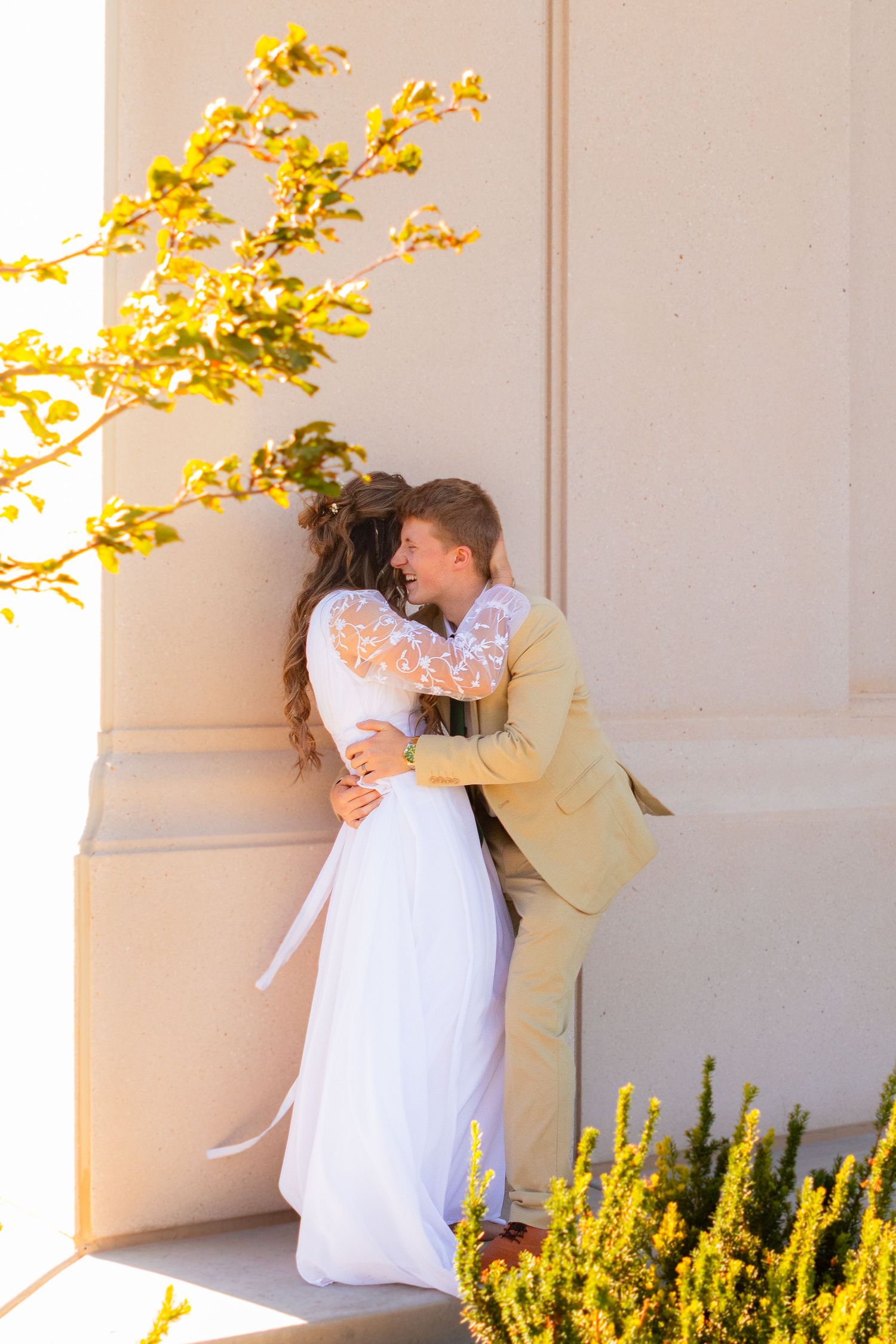 Bride and groom embracing near a stone pillar, surrounded by yellow flowers and green foliage on a sunny wedding day.