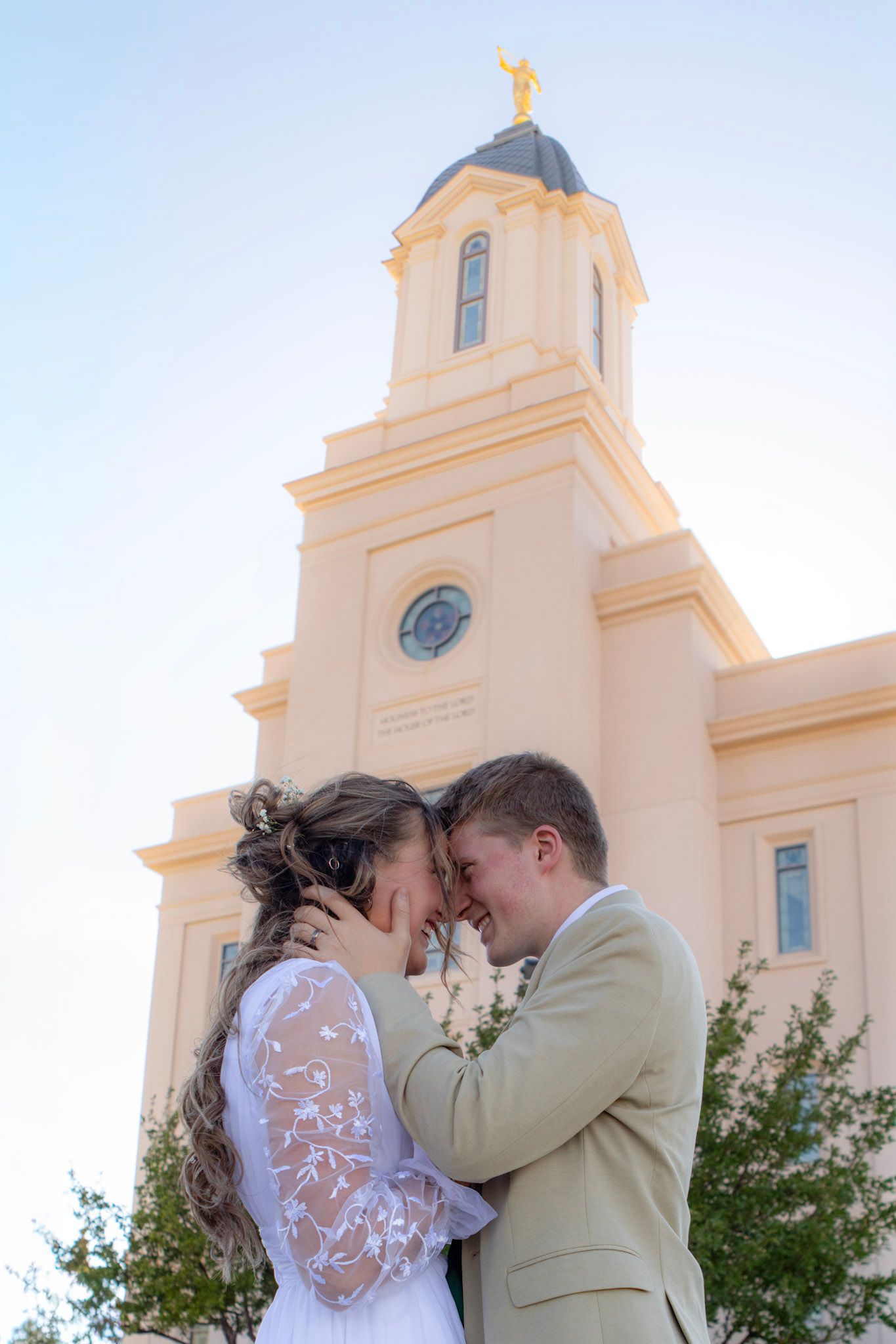 Bride and groom touching foreheads romantically in front of an LDS temple spire with golden angel Moroni on their wedding day.