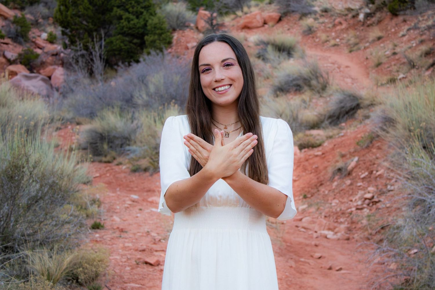 Smiling young woman in white dress clasping hands on red rock desert trail backdrop.