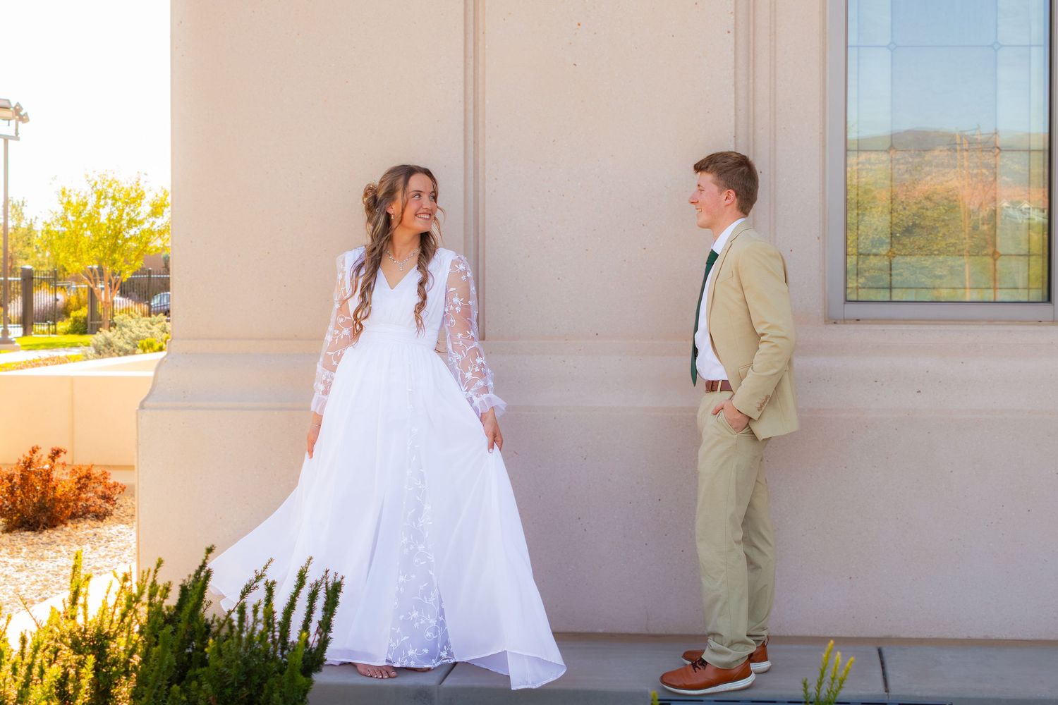 Bride in white lace gown and groom in tan suit share a joyful moment outside a building on their wedding day.