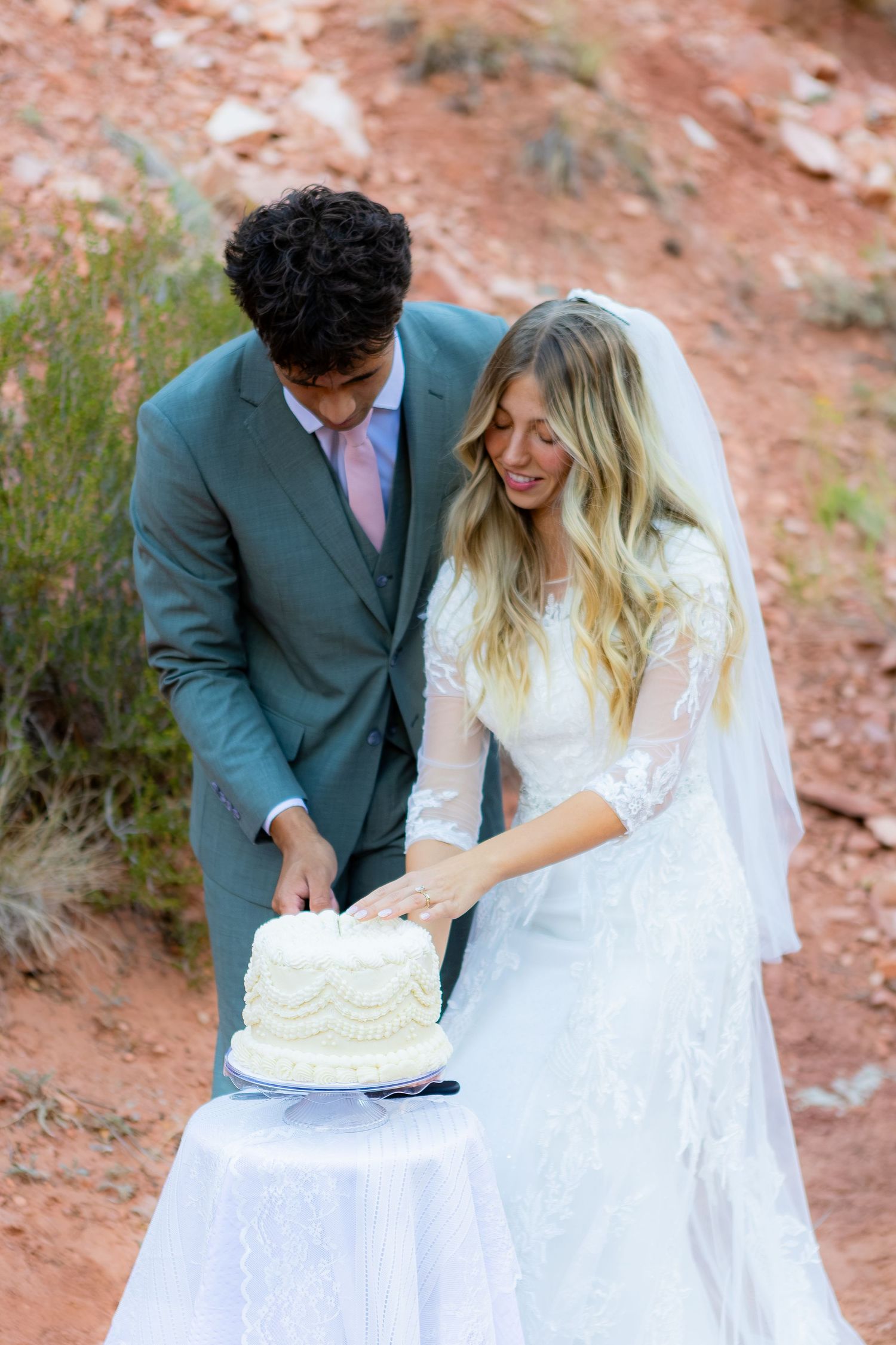 Bride and groom cutting white wedding cake outdoors in red rock desert setting, bride in lace dress with veil.