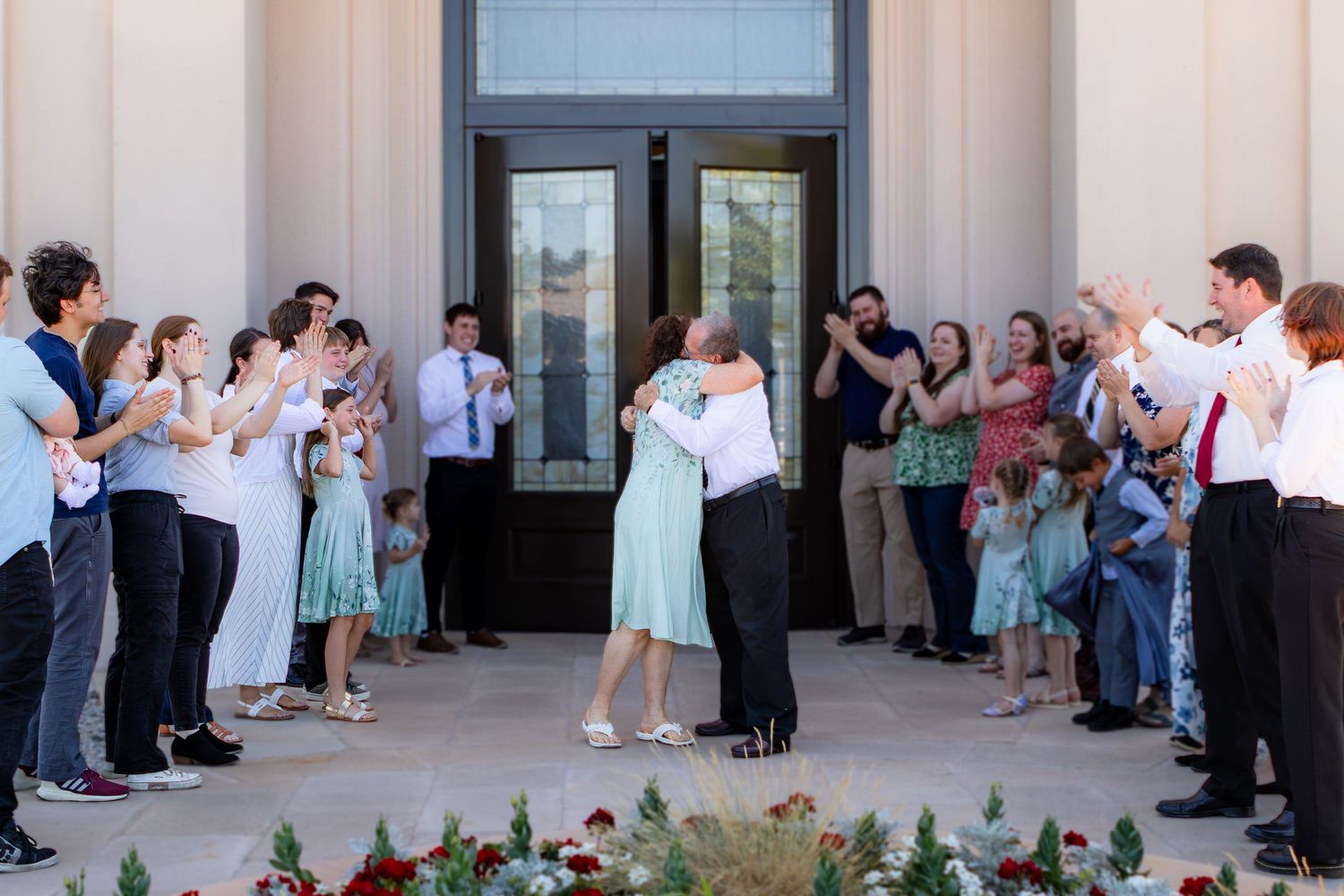 Couple shares first dance outside wedding venue surrounded by cheering guests in formal attire on sunny day.