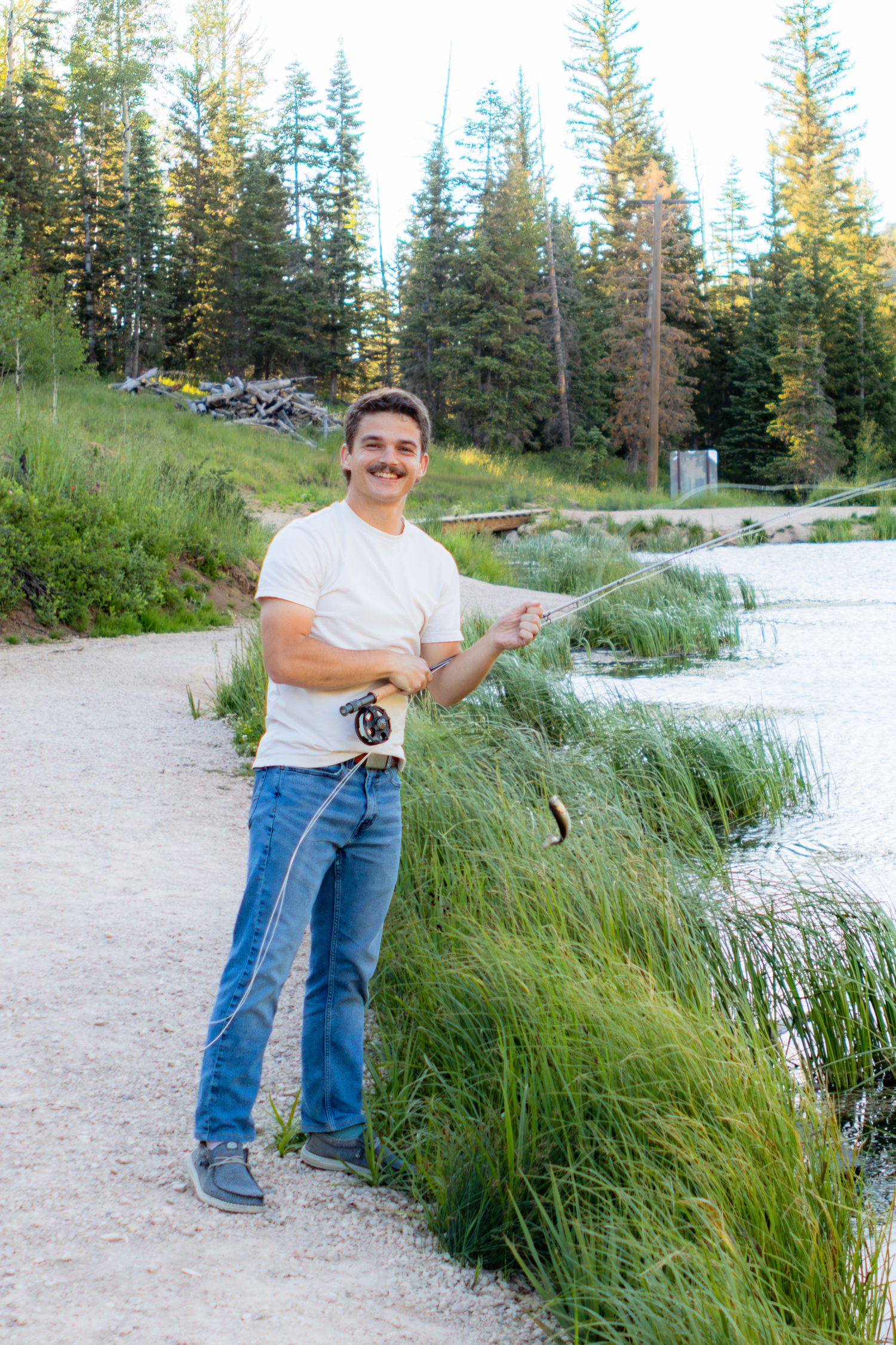 Man with mustache fly fishing by a mountain lake surrounded by tall green grass and pine trees.