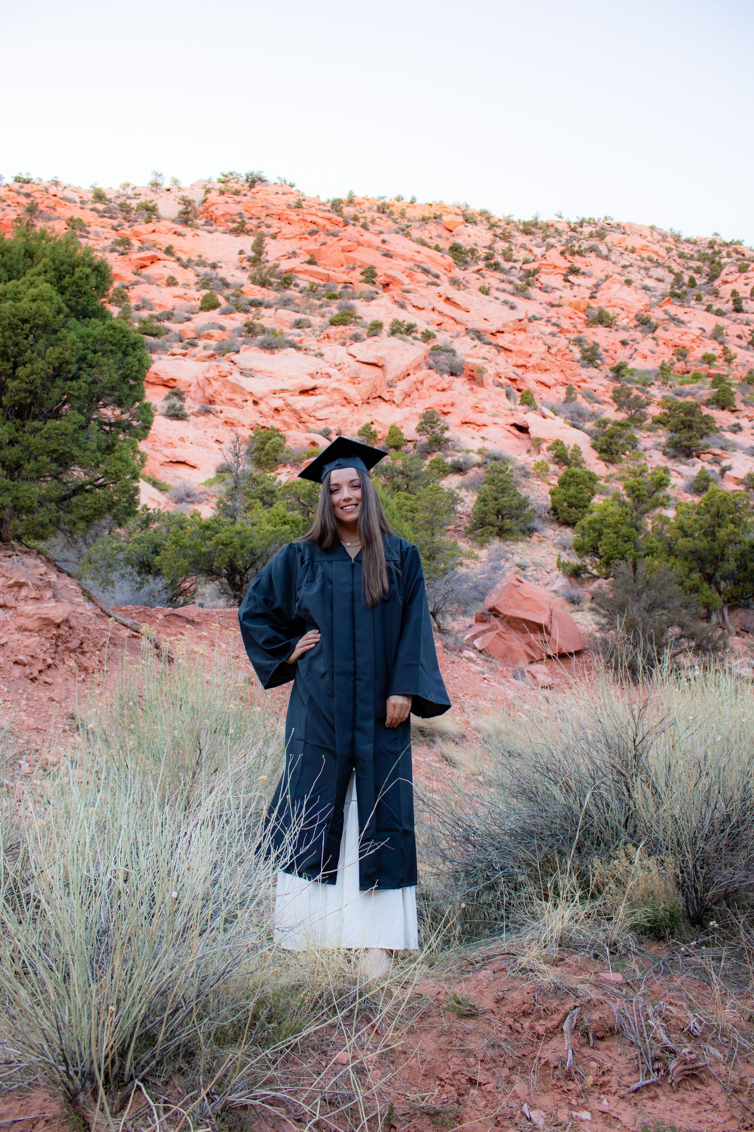 Female graduate in black cap and gown standing confidently in desert brush with red rock hills.