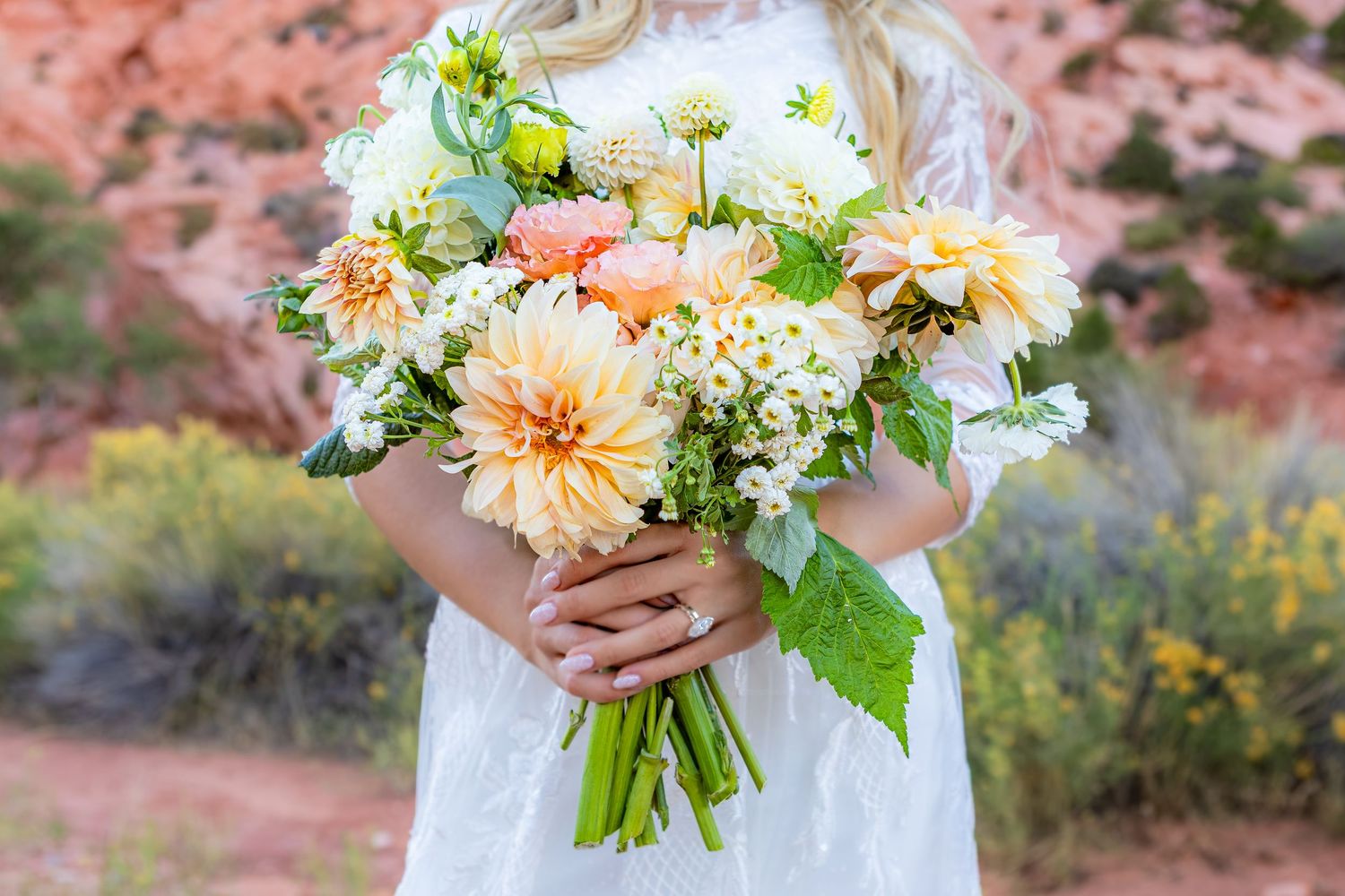 Bride holding a peach and white dahlia wedding bouquet with greenery outdoors in a desert landscape.