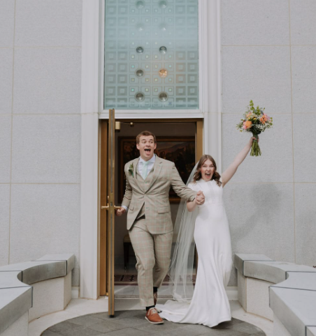 Joyful newlyweds exit church doors, bride in white gown holding colorful bouquet, groom in beige suit, celebrating marriage.