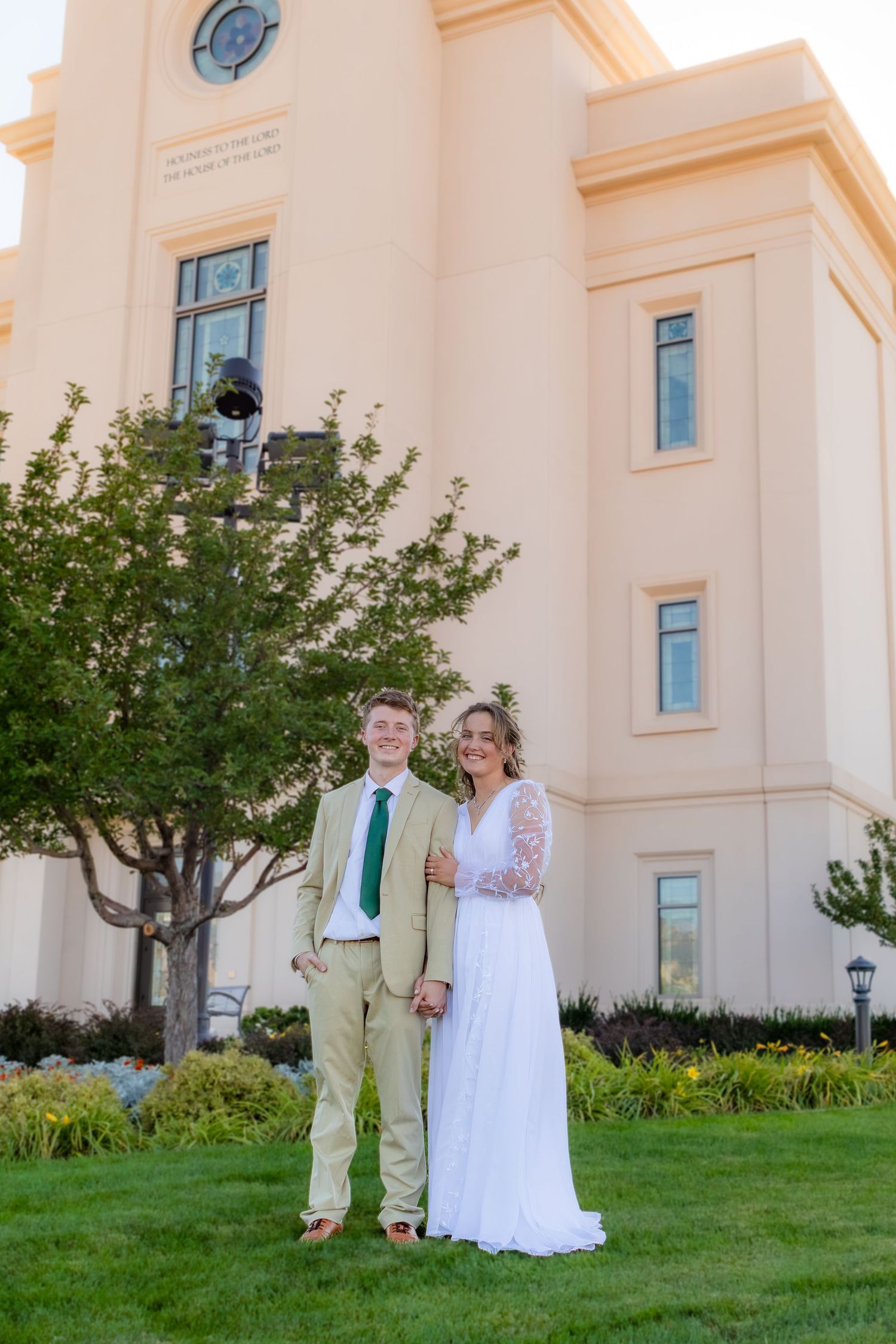 Newlywed couple posing in front of an LDS temple, groom in tan suit, bride in white lace wedding dress.