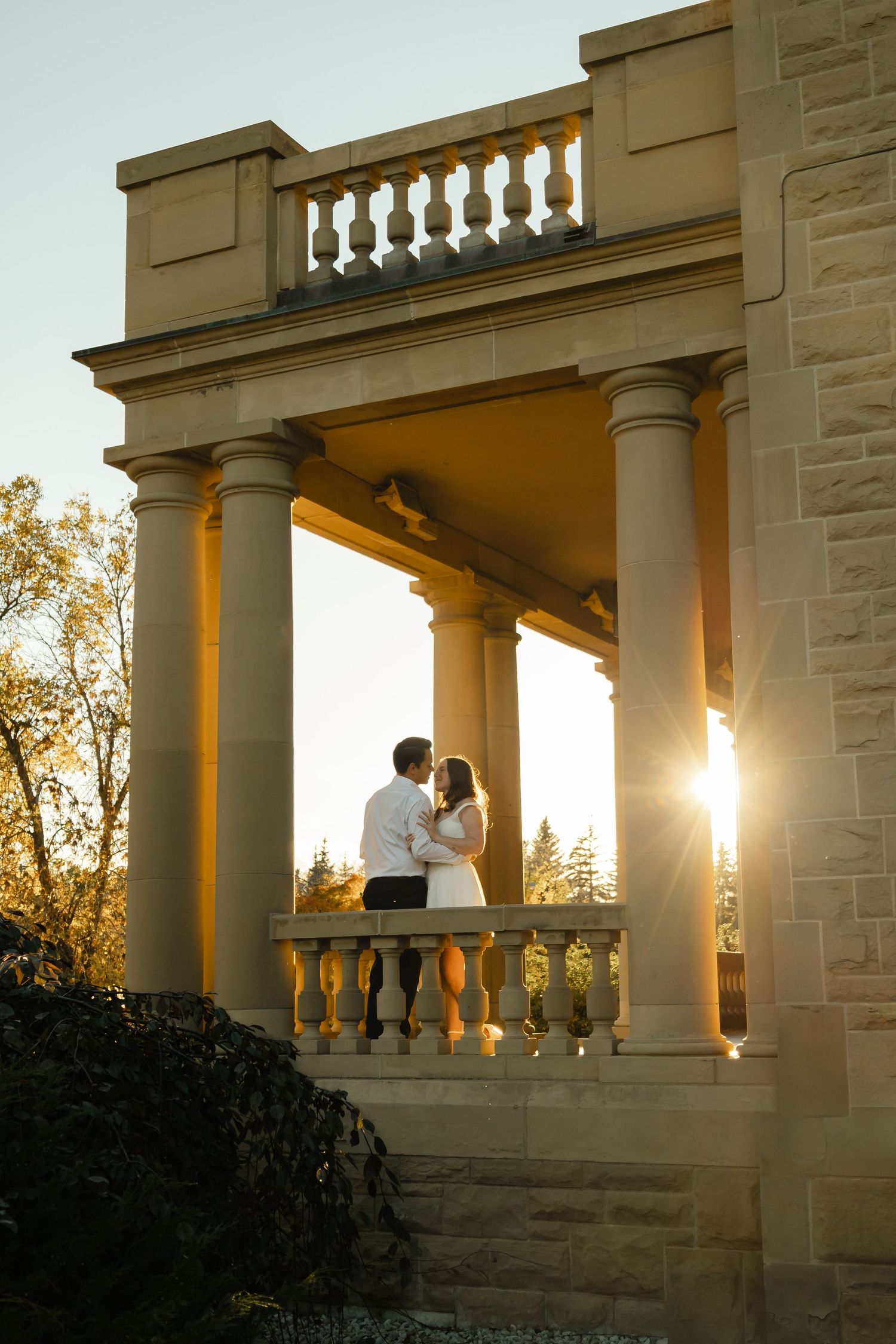An engaged couple standing together on the balcony of Government House as the sun sets in Edmonton, Alberta.