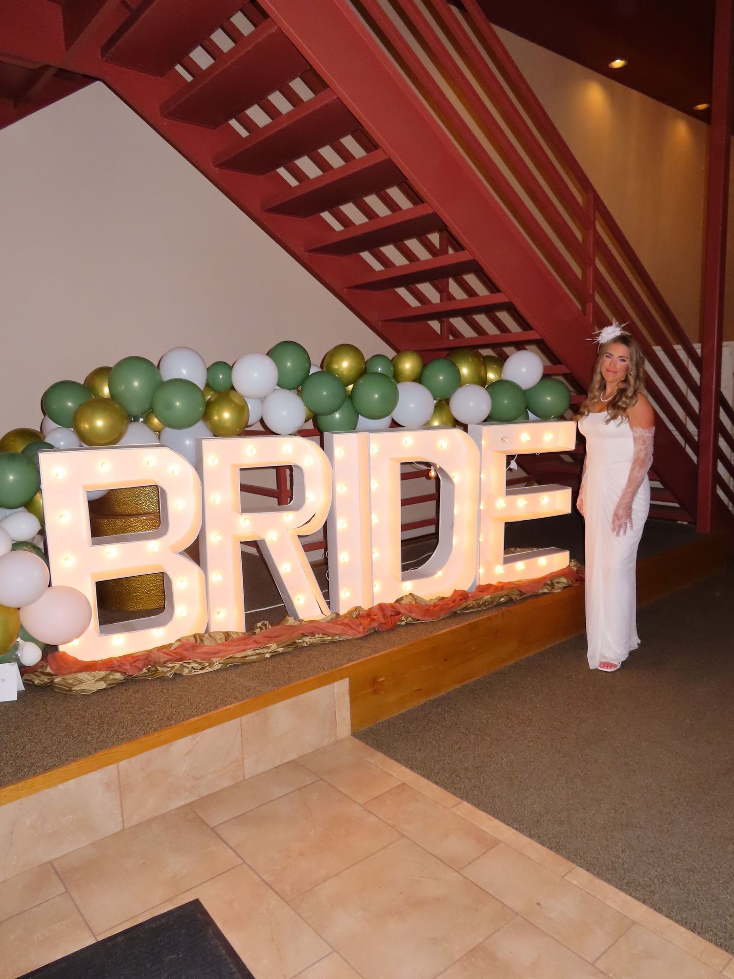 Bride in white dress posing next to large illuminated BRIDE letters with green balloon arch.
