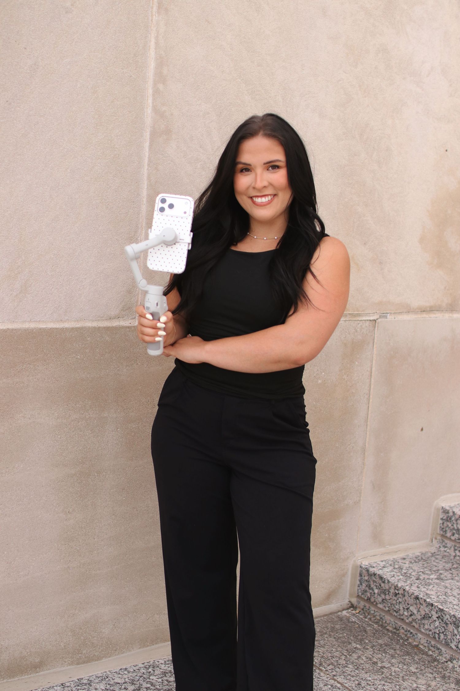 Woman in black outfit smiling while holding smartphone on gimbal stabilizer outdoors.
